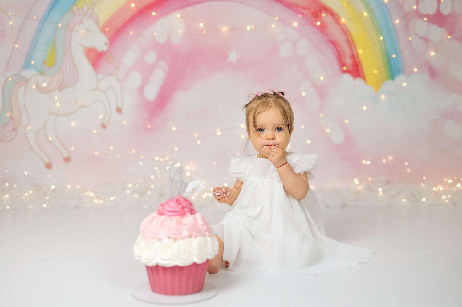 Baby girl in white dress sits beside a pink cupcake smash cake with unicorn rainbow backdrop and fairy lights.