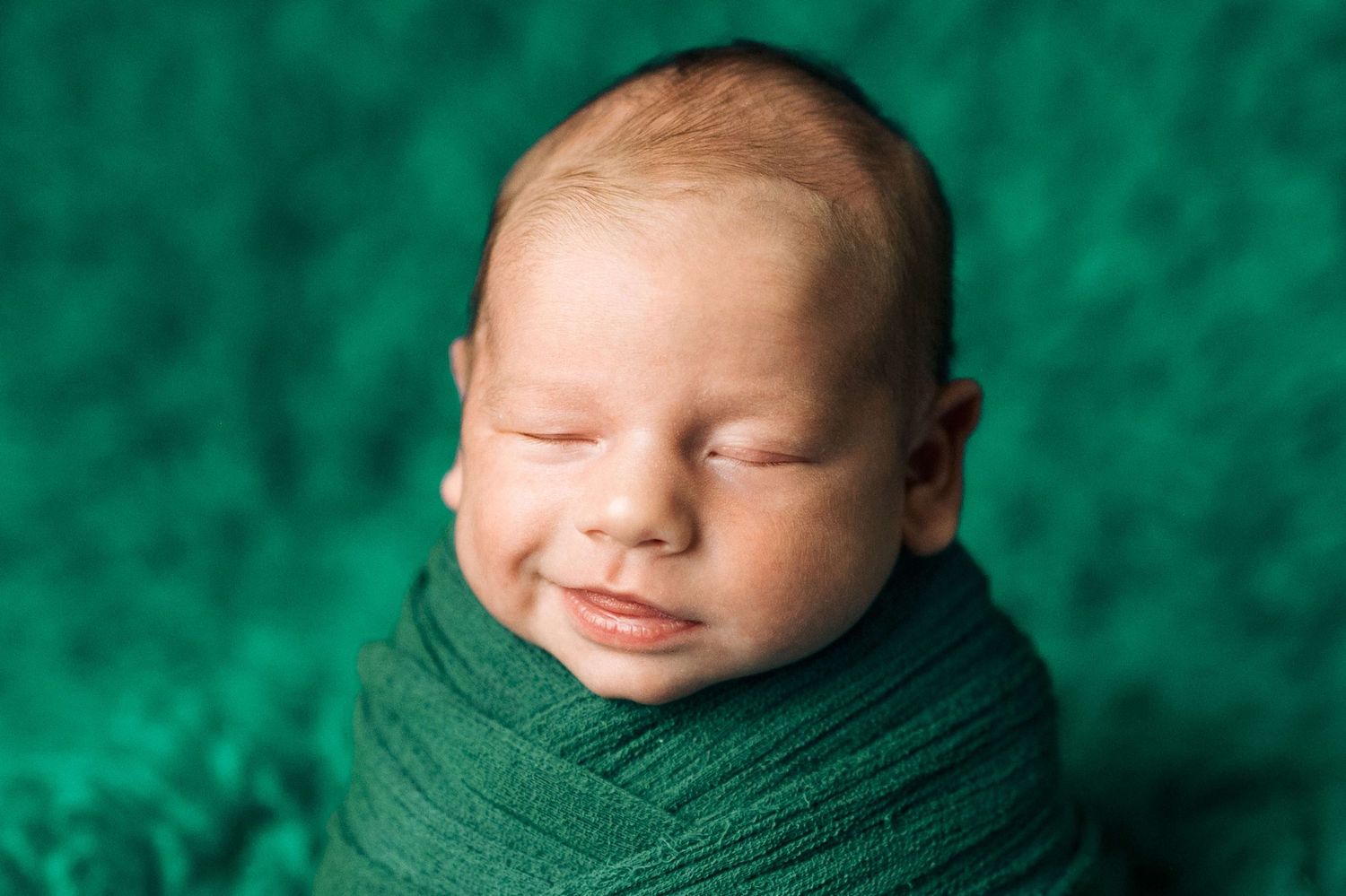 Sleeping newborn baby swaddled in green wrap on teal fuzzy blanket, peaceful smile on face.