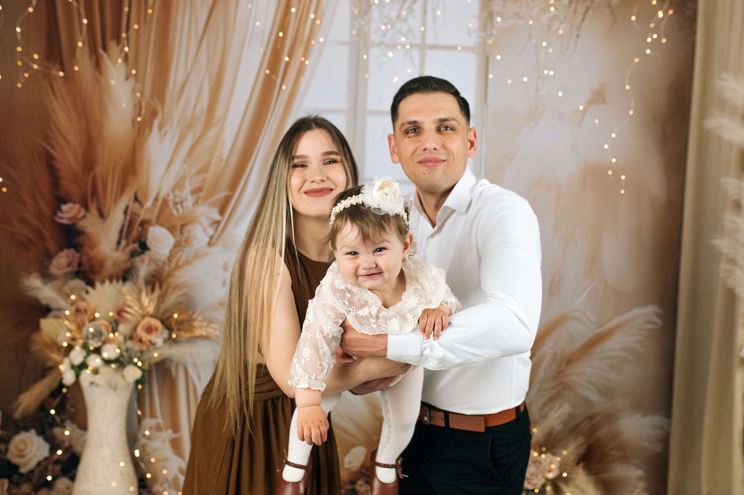 A smiling family with a mother, father, and baby girl posing in a warm, elegant studio with floral decor.