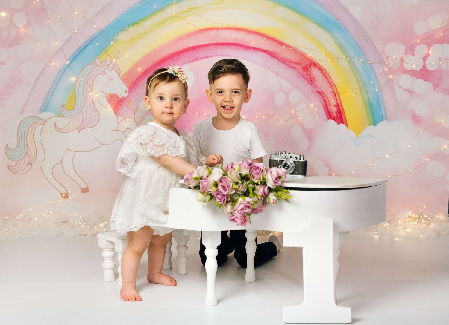 Two young children pose by a white mini piano with flowers, in front of a colorful rainbow unicorn backdrop.