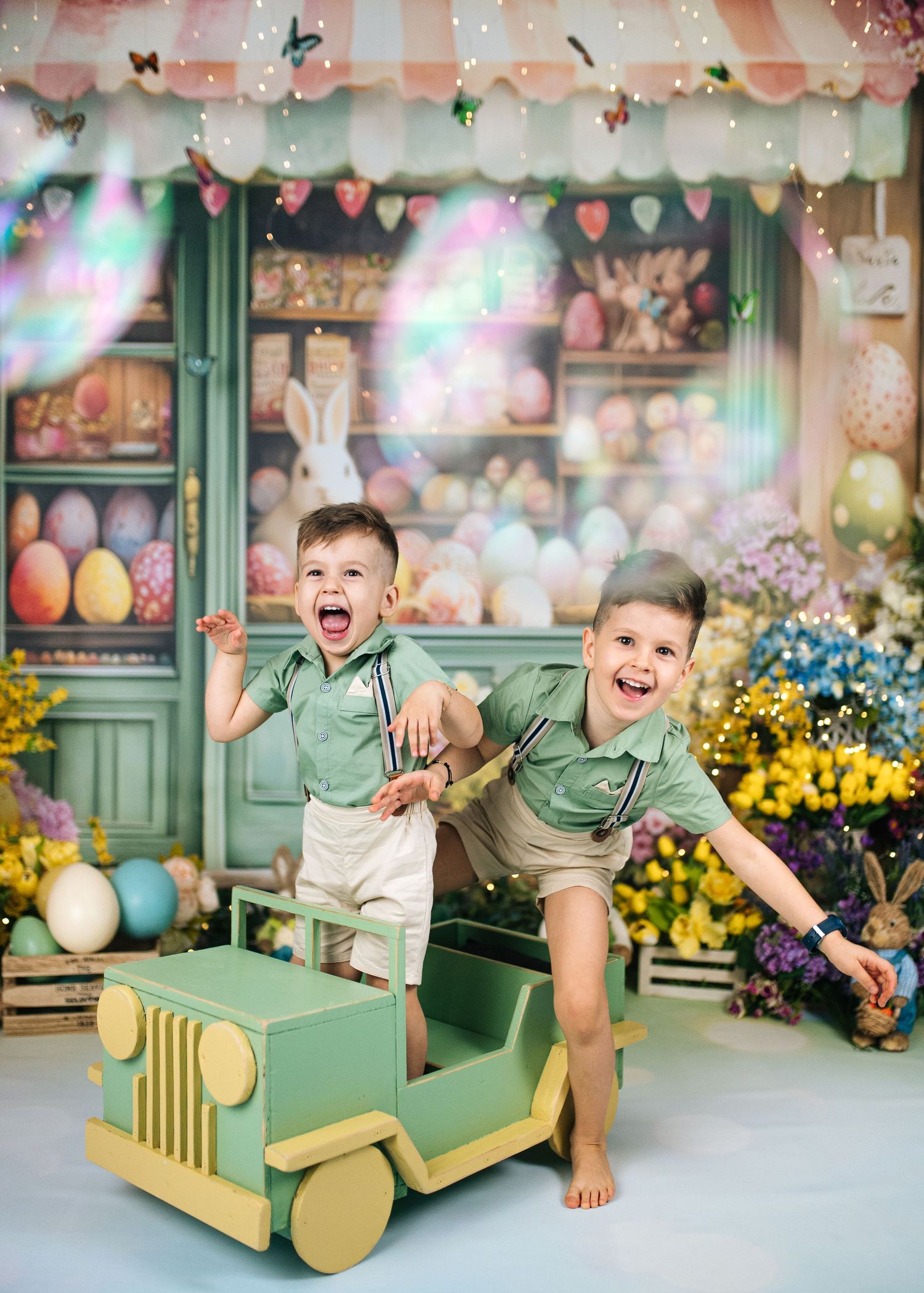 Two boys in matching green shirts laugh and play in a wooden toy jeep amid a colorful Easter-themed backdrop.