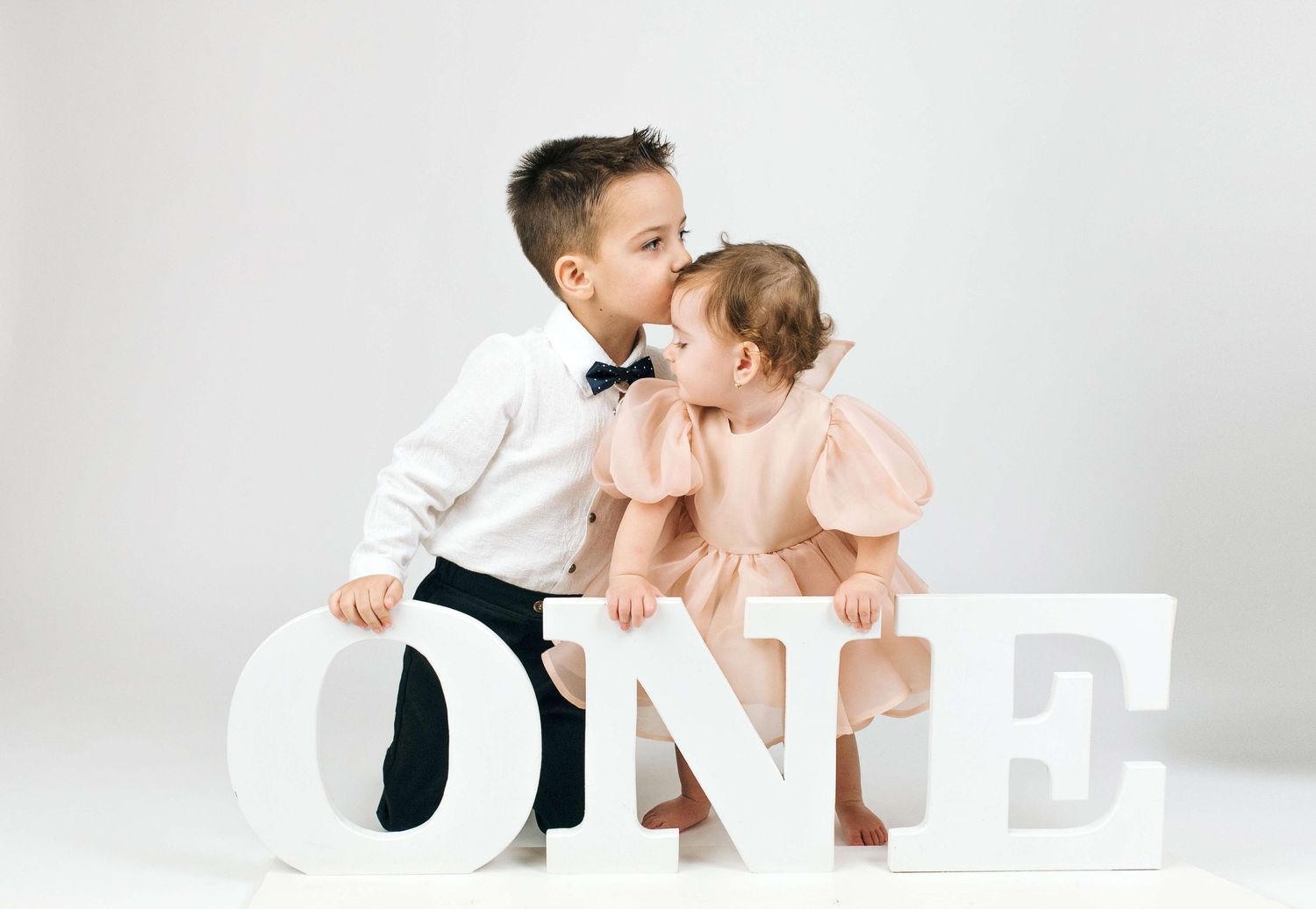A young boy in a bow tie kisses a baby girl in a pink dress beside large white ONE letters.