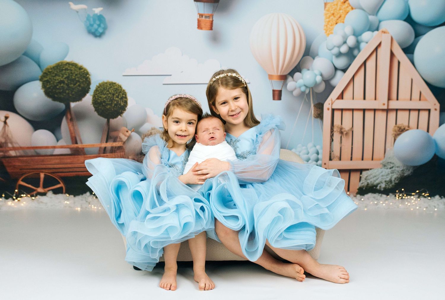 Two girls in blue dresses pose with a newborn baby against a whimsical hot air balloon backdrop.