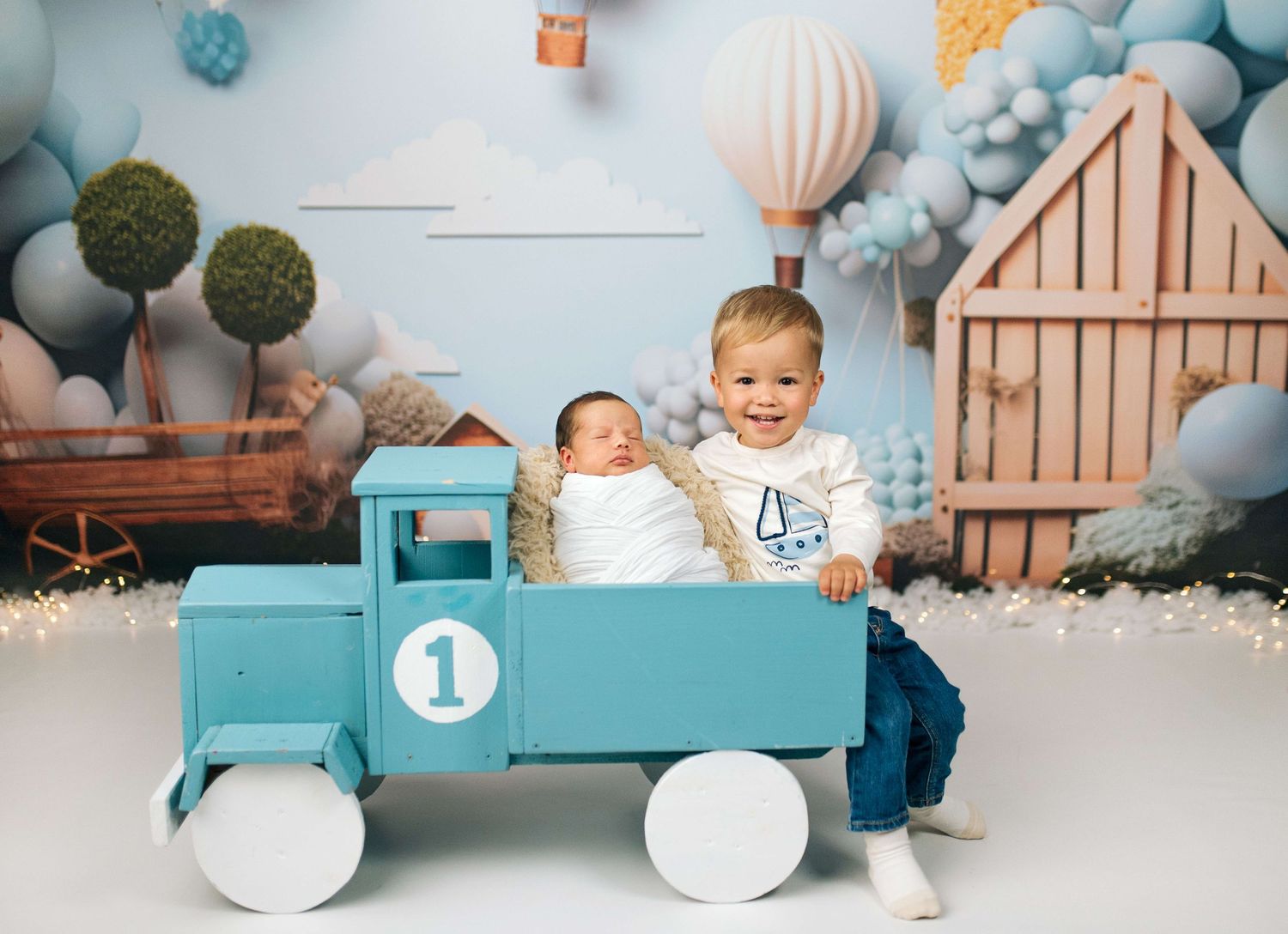 Two boys pose in a teal toy truck with hot air balloon backdrop in a whimsical studio photo shoot.