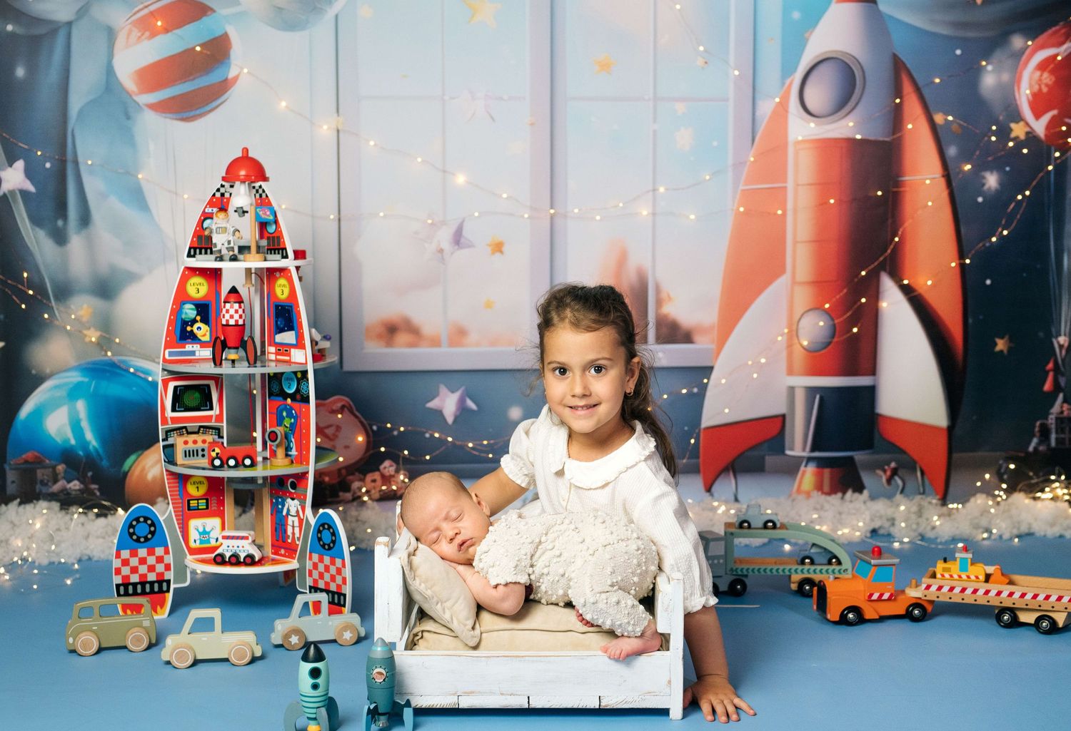 Young girl holds sleeping newborn baby on white bed surrounded by space-themed toys and rocket backdrop.