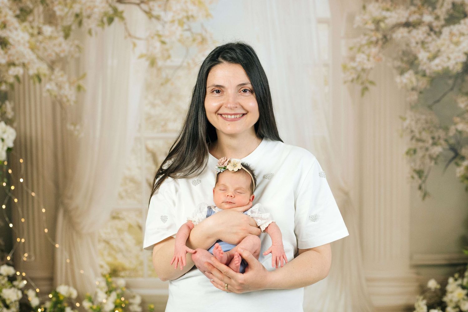 Smiling young mother in white dress holds sleeping newborn baby with floral headband in elegant studio setting.