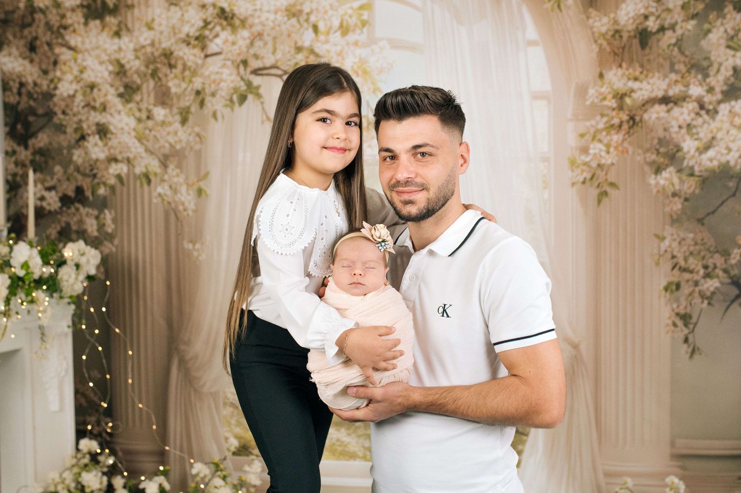 A young family poses with their newborn baby in a floral studio setting, wearing white and black outfits.