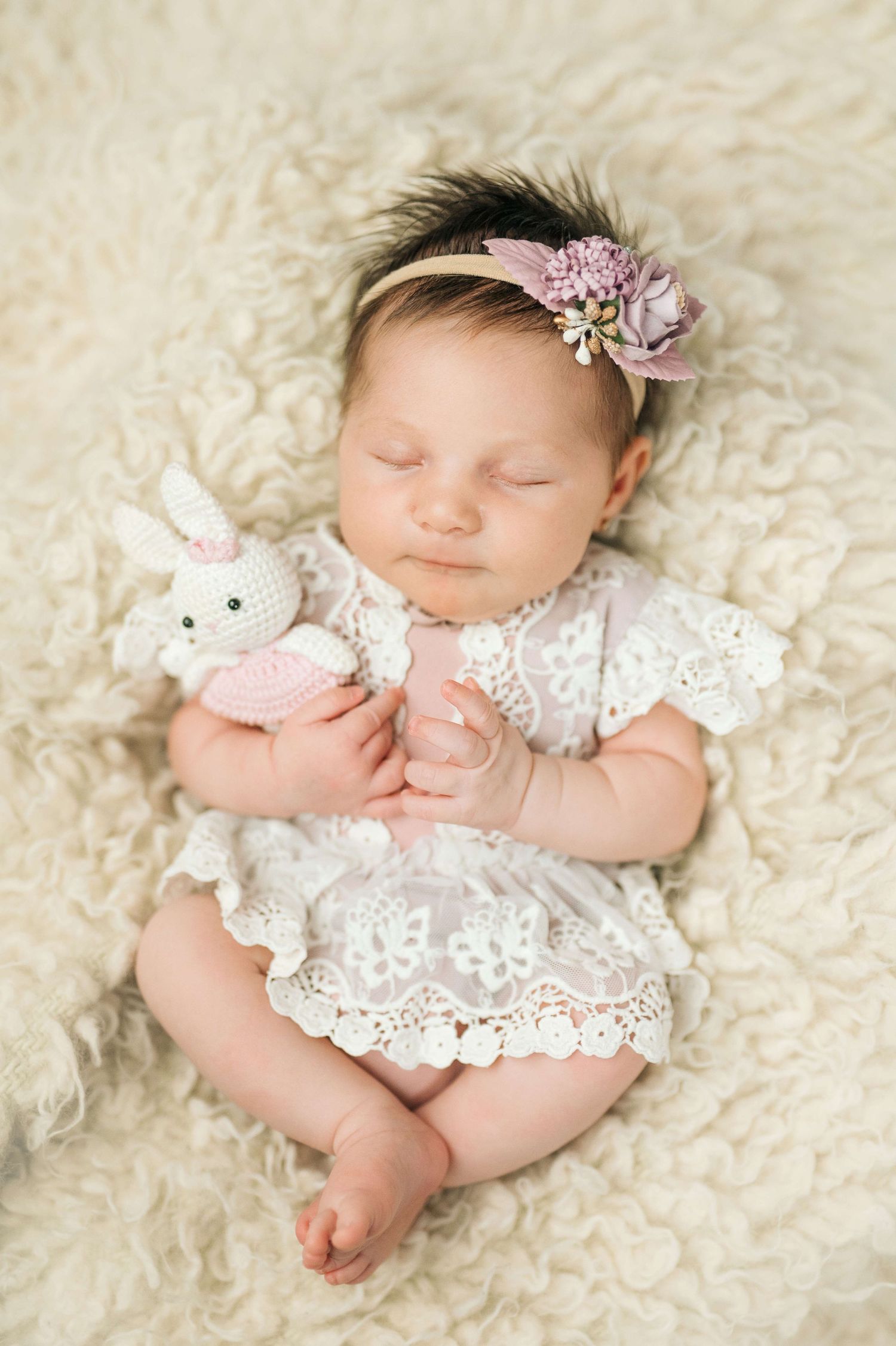 Sleeping newborn baby girl in white lace dress holding crochet bunny toy, wearing floral headband on fluffy cream blanket.