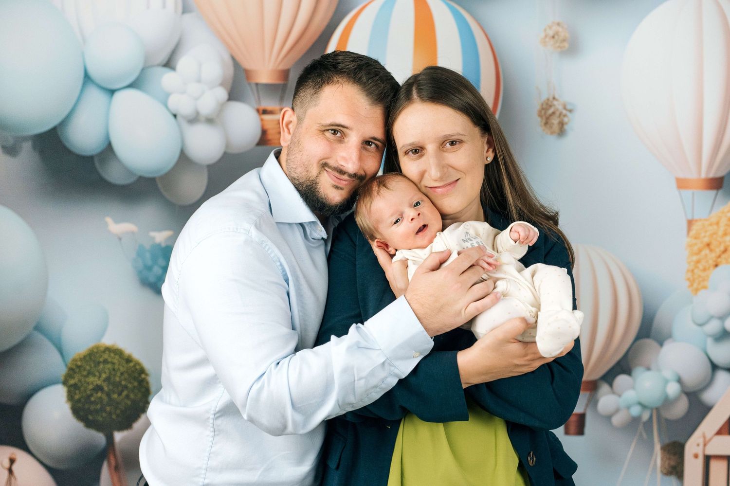 Happy young family with newborn baby posing in front of colorful hot air balloon backdrop in professional photo studio.