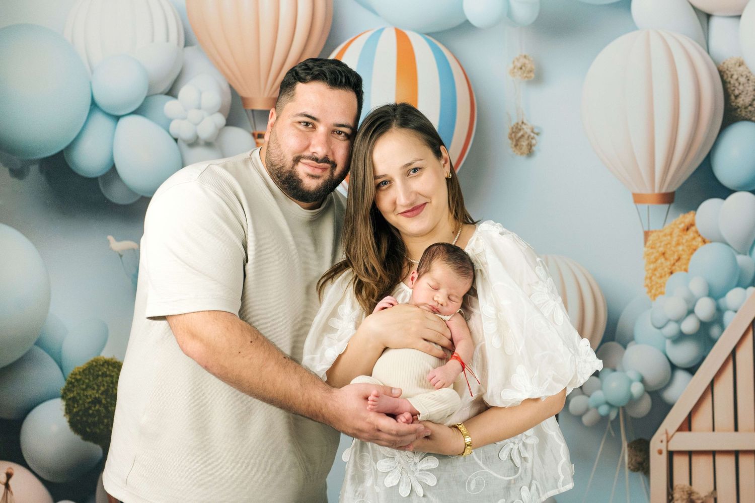 Happy couple holding their newborn baby in front of a pastel blue balloon and hot air balloon backdrop.