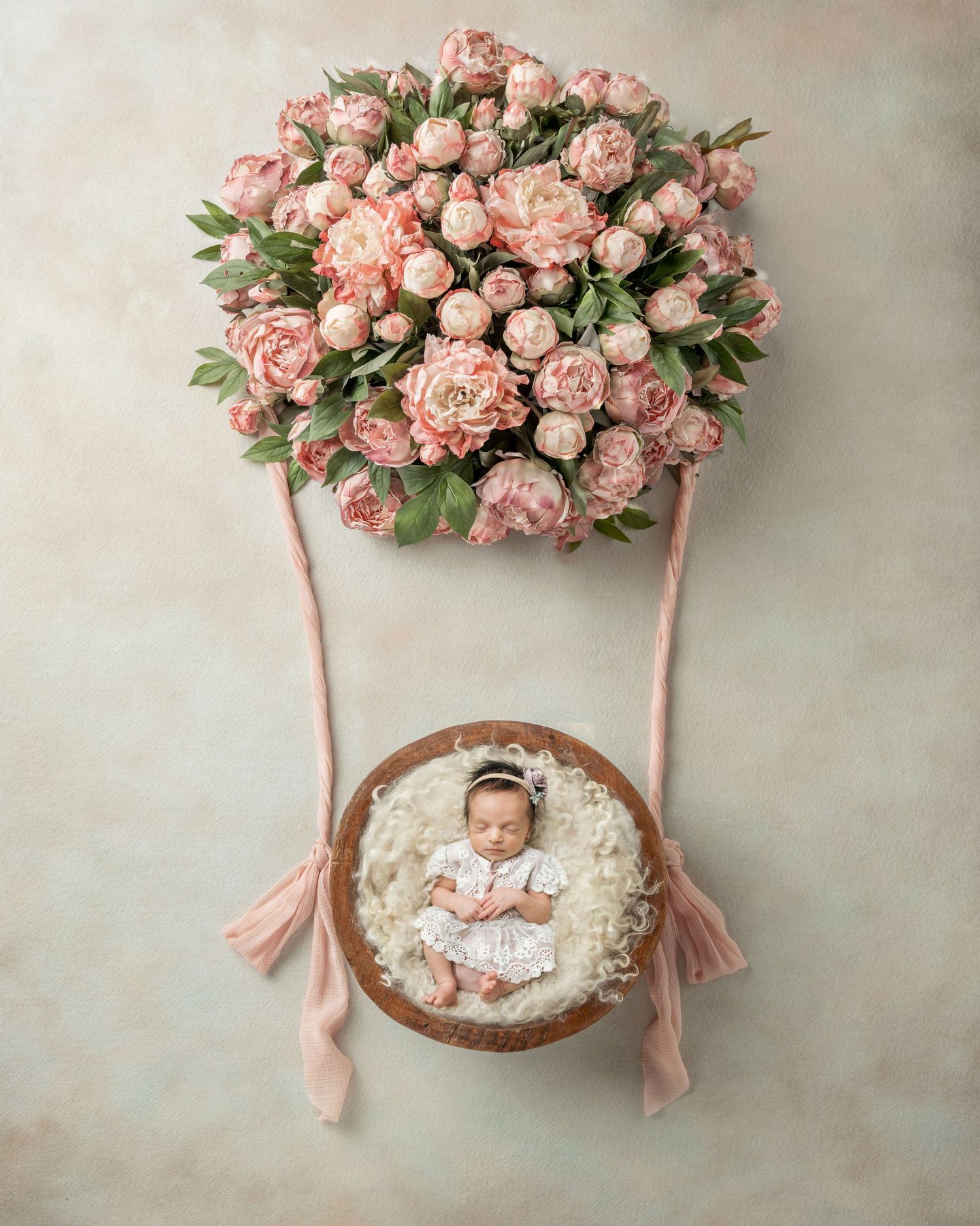 Newborn baby girl sleeping in a wooden bowl basket beneath a pink peony hot air balloon arrangement.