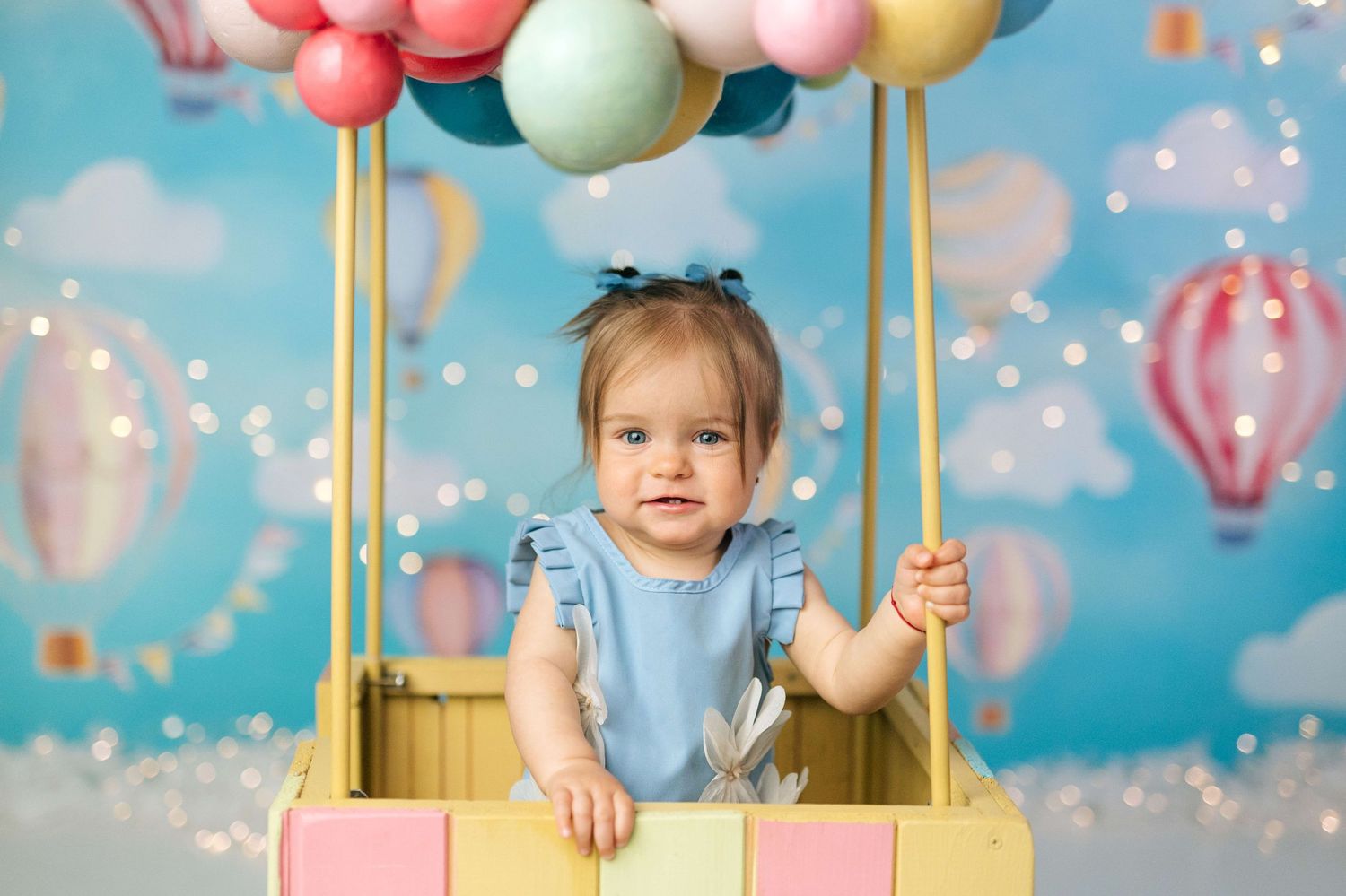 A baby girl in a blue dress sits in a colorful hot air balloon prop with pastel balloons overhead.