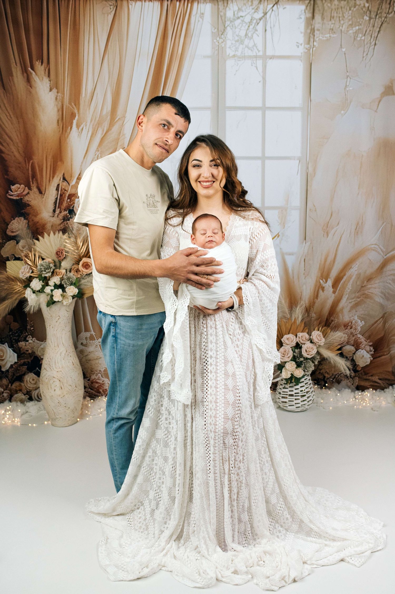 A happy couple in a boho studio holding their newborn baby, woman in white lace gown with floral decor backdrop.