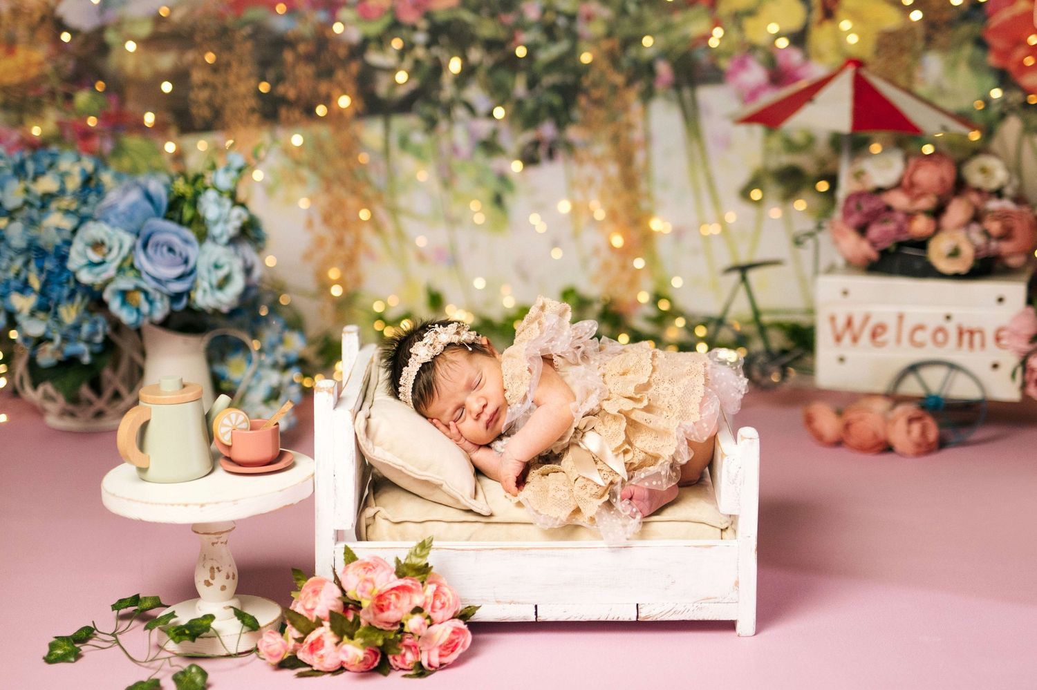 Newborn baby sleeping peacefully in a tiny white bed surrounded by flowers and fairy lights in a whimsical welcome setup.
