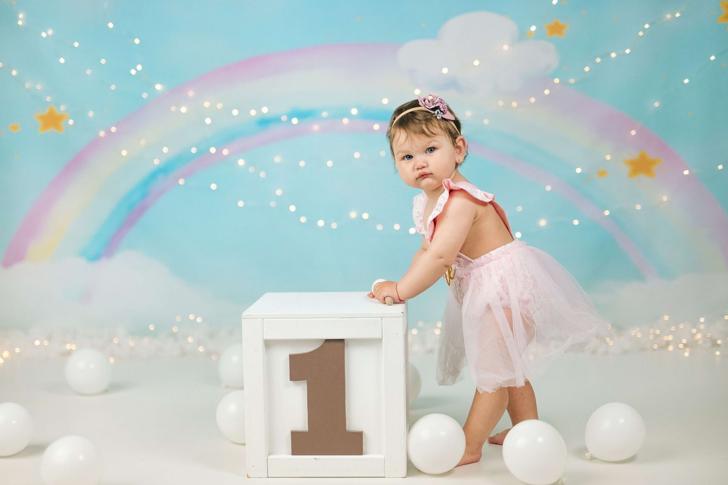 Baby girl in pink tutu celebrates first birthday with rainbow backdrop, white balloons, and number one block.