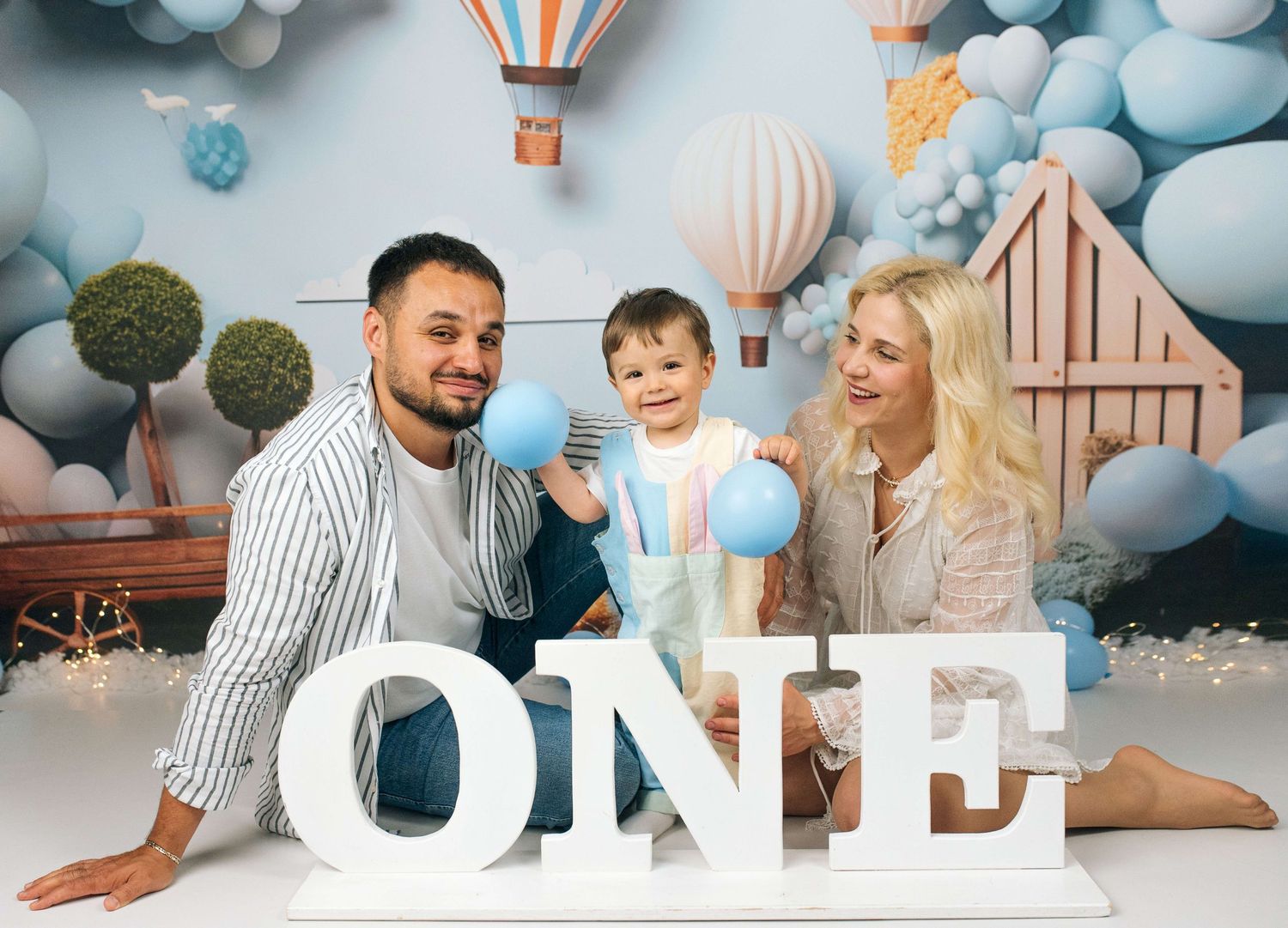 Family of three celebrating baby’s first birthday with blue balloons and ONE letters in a festive photo studio.