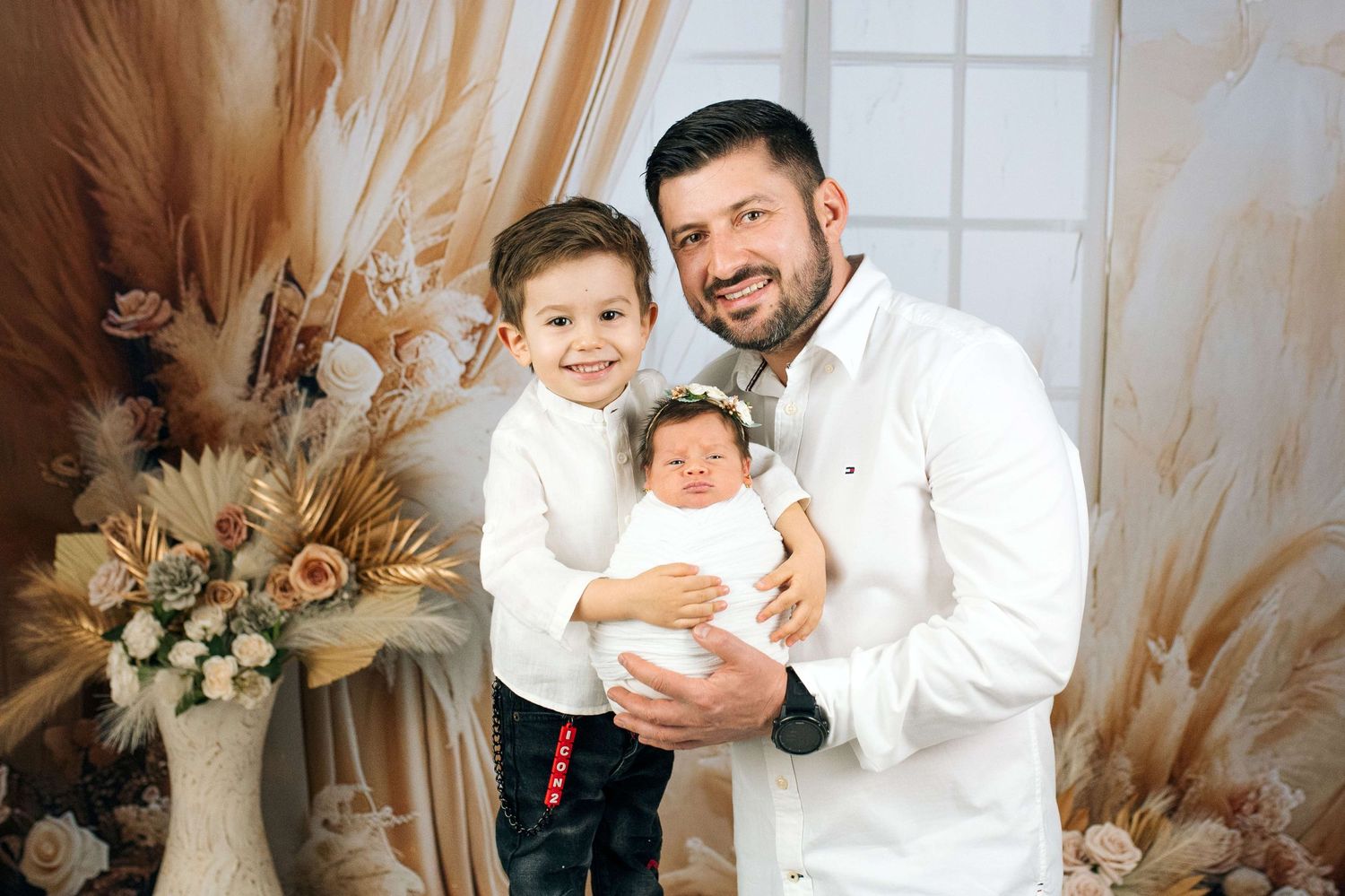 Father smiling with young son and newborn baby in arms, posed in a warm floral decorated photo studio.