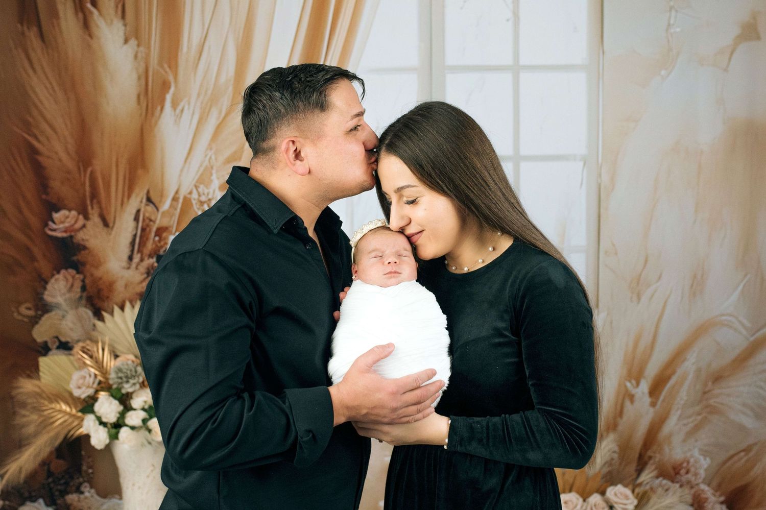 A couple in matching black outfits tenderly kiss their newborn baby wrapped in white during a studio photo session.