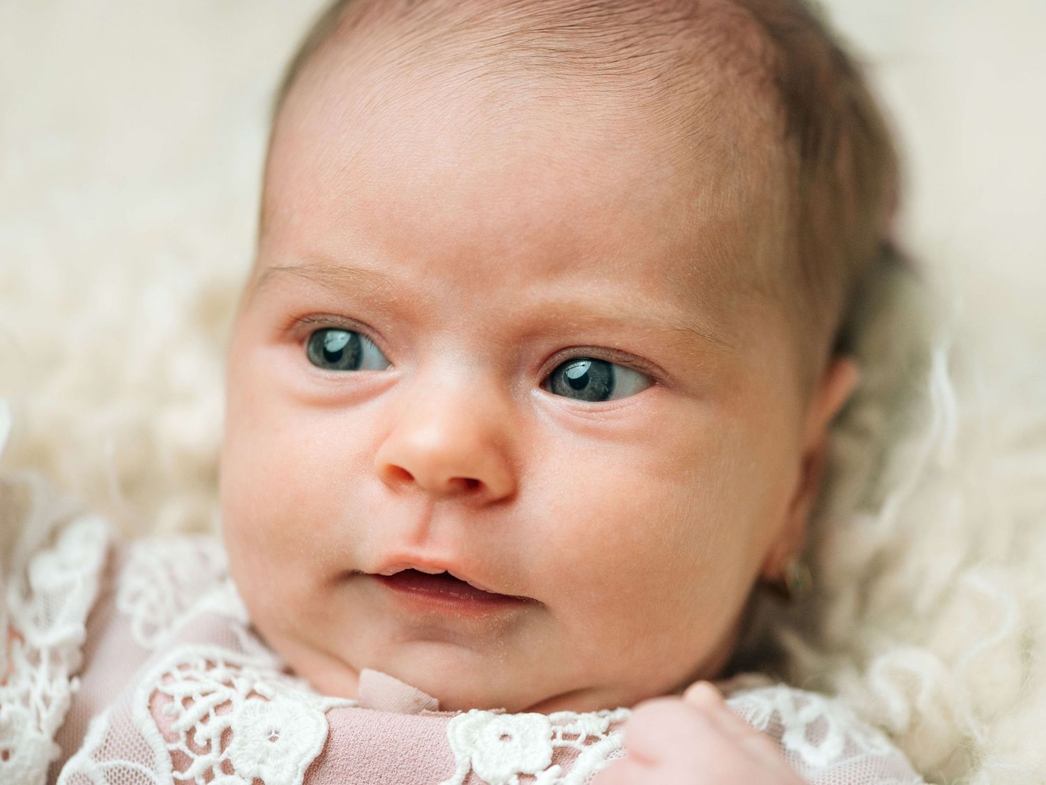 Close-up portrait of a newborn baby with blue eyes wearing a white lace outfit, lying on a soft cream blanket.