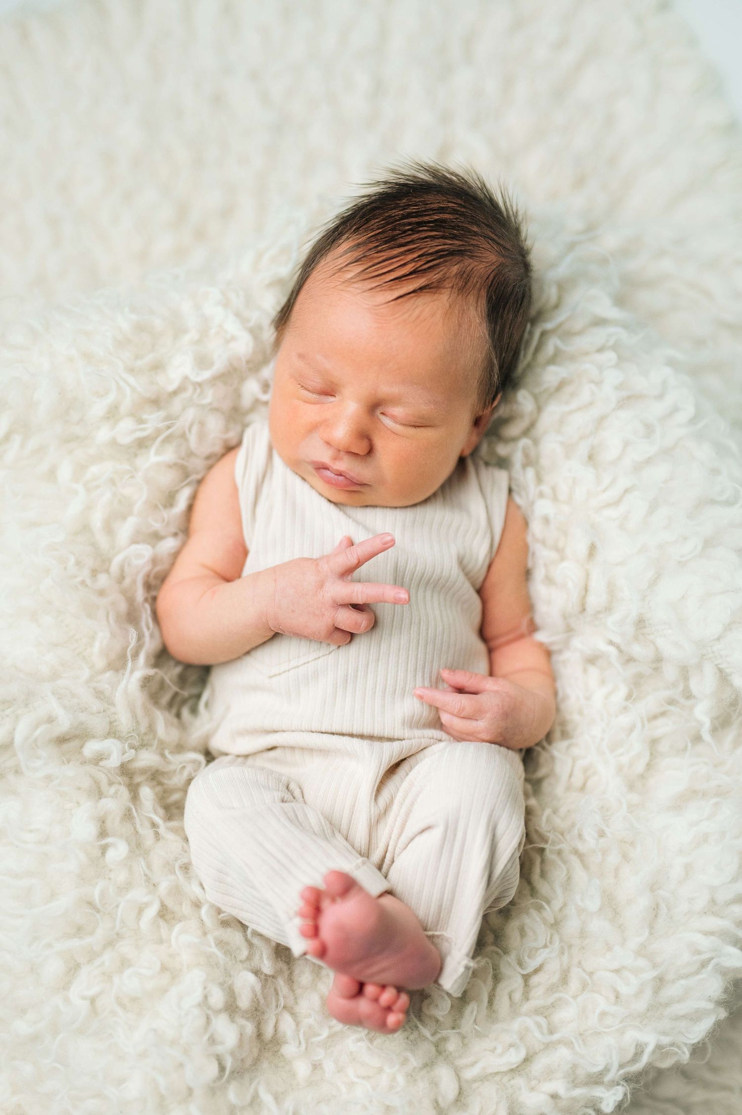 Peaceful newborn baby sleeping on a fluffy white blanket wearing a cream ribbed outfit with dark hair.