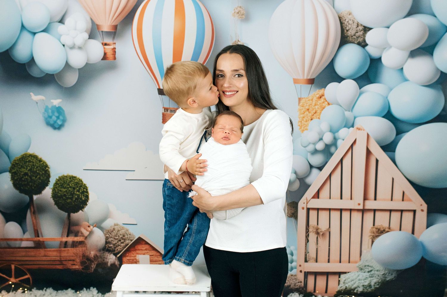 A smiling mother holds a newborn baby while her toddler stands beside her, surrounded by pastel balloons and hot air balloon decor.