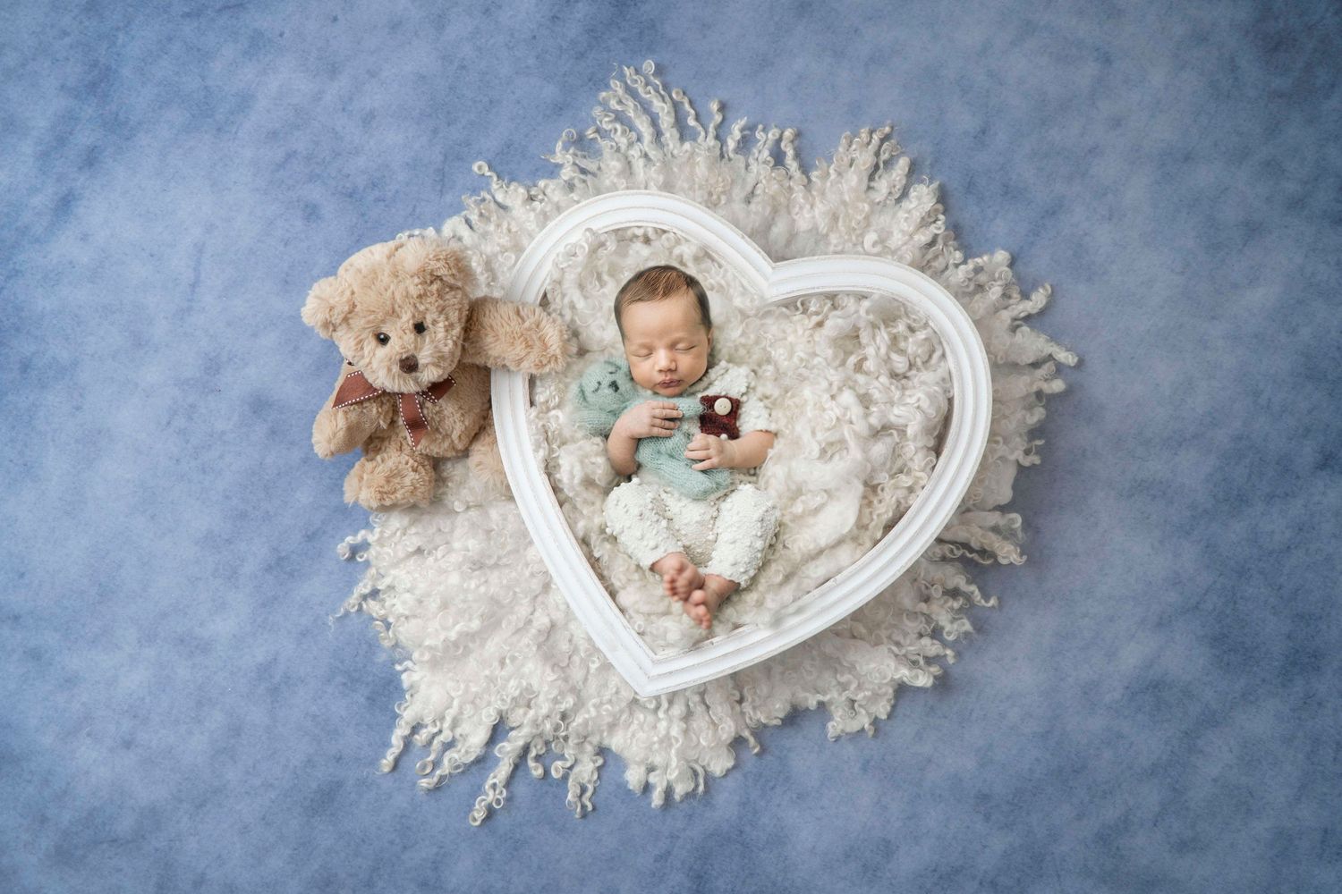 Newborn baby sleeping in white heart-shaped bowl with teddy bear on blue background.