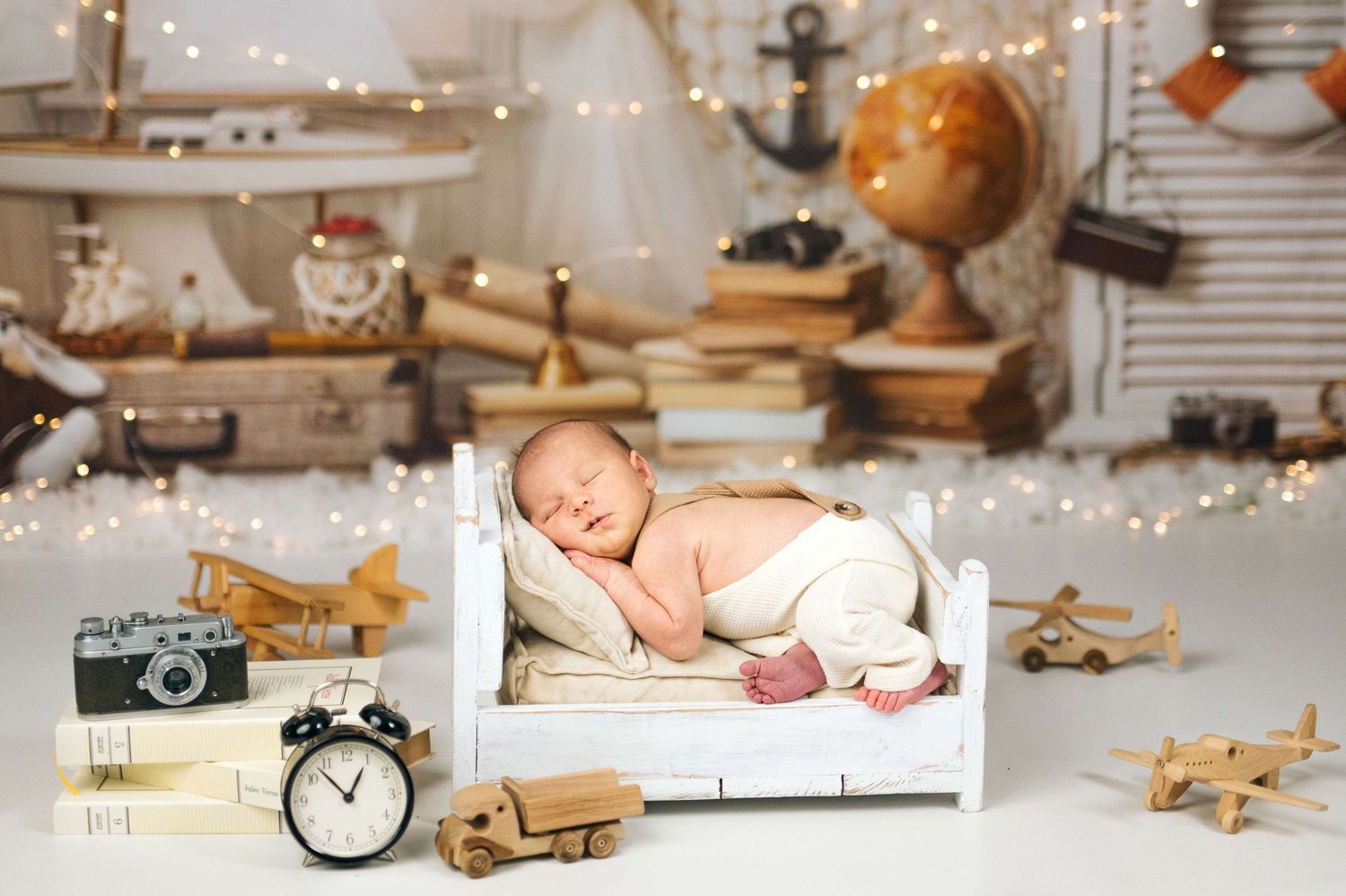 Newborn baby sleeping peacefully on a tiny white bed surrounded by wooden toys, books, globe, and fairy lights.