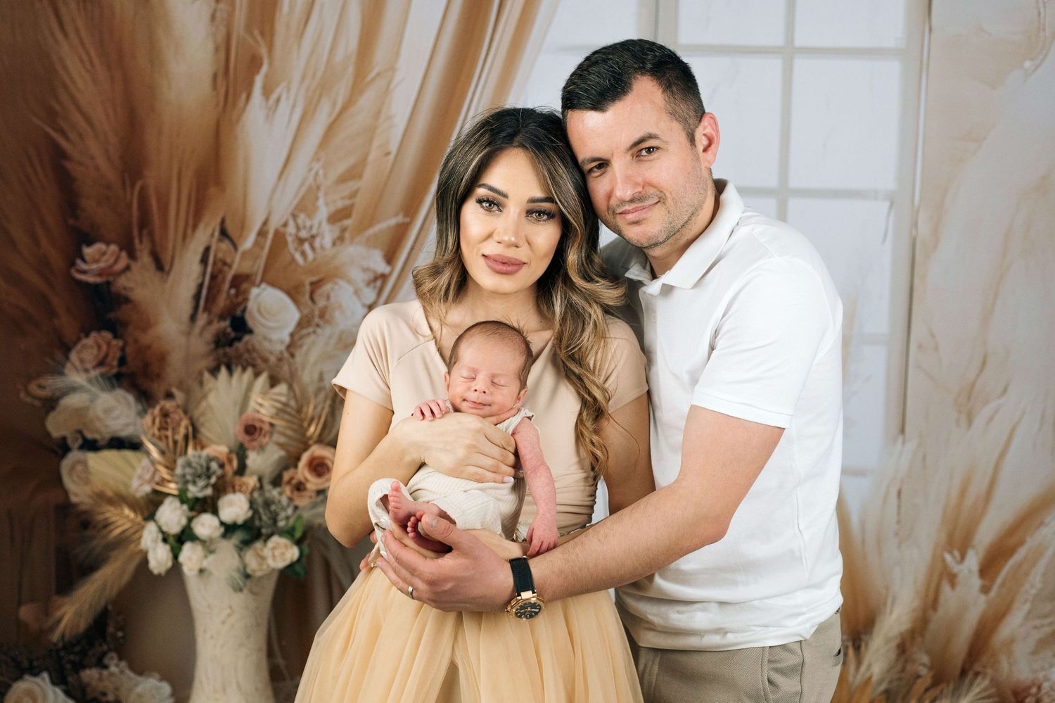 A happy couple in a boho studio setting poses with their newborn baby, dressed in elegant neutral tones.