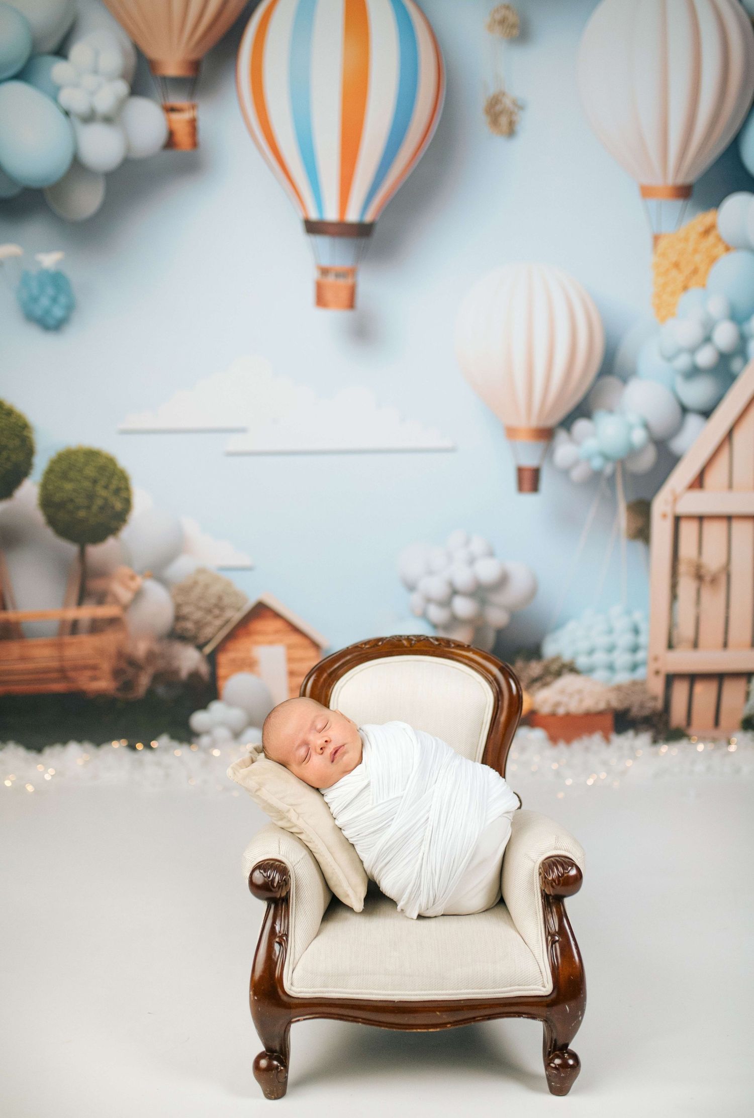 Newborn baby swaddled in white, sleeping peacefully in a miniature chair with hot air balloon backdrop.