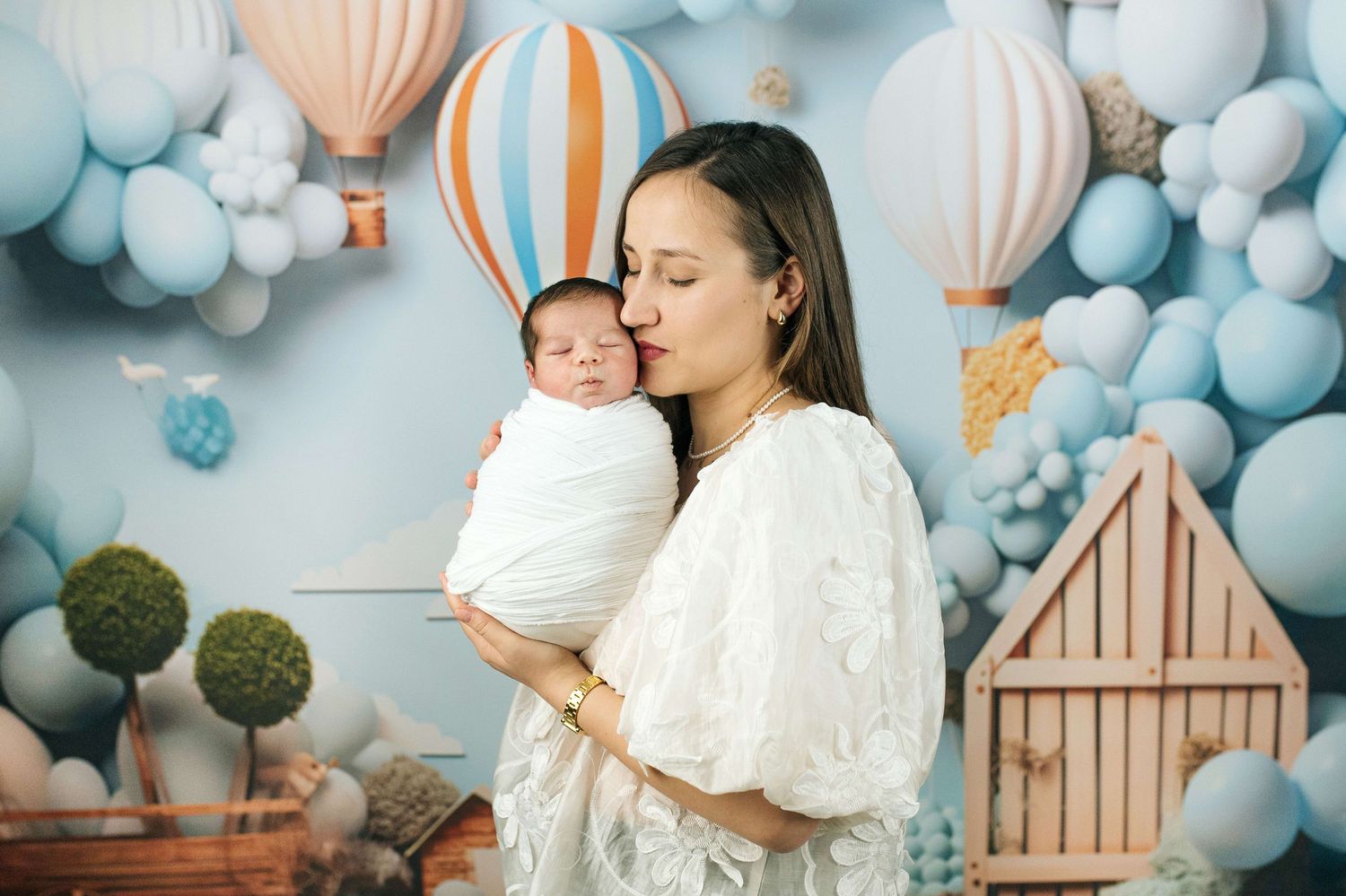 Mother in white dress tenderly kisses swaddled newborn baby against blue balloon and hot air balloon backdrop.