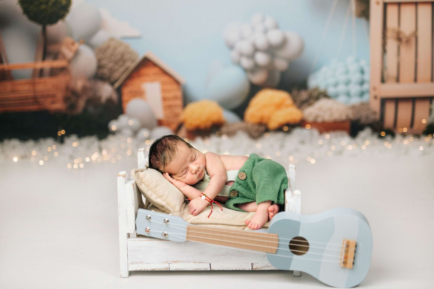 Newborn baby sleeping peacefully on a tiny white bed with a small blue guitar, surrounded by a whimsical nursery backdrop.