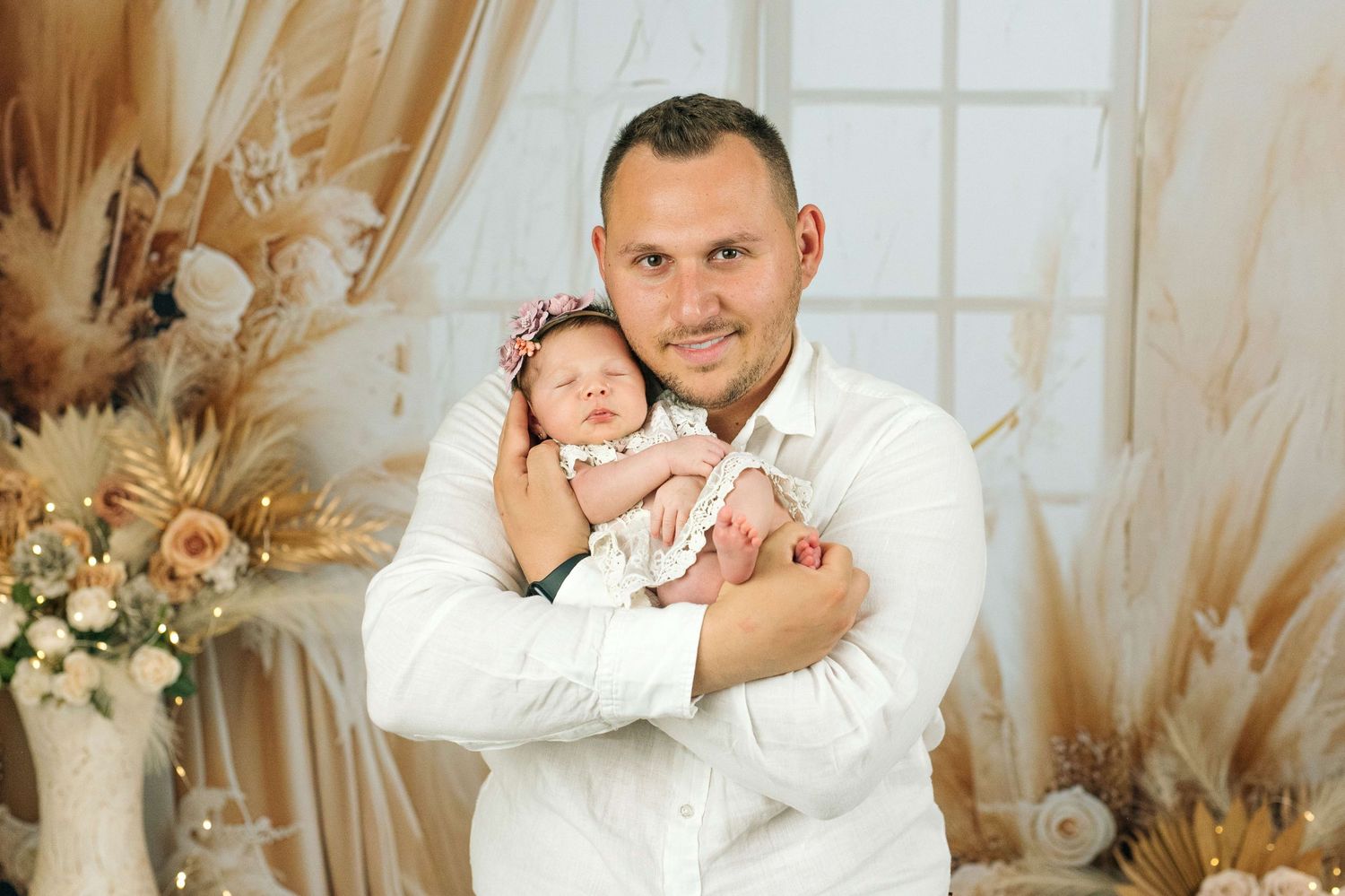 A proud father in a white shirt holds his newborn baby girl in a soft, floral boho-styled photography studio.