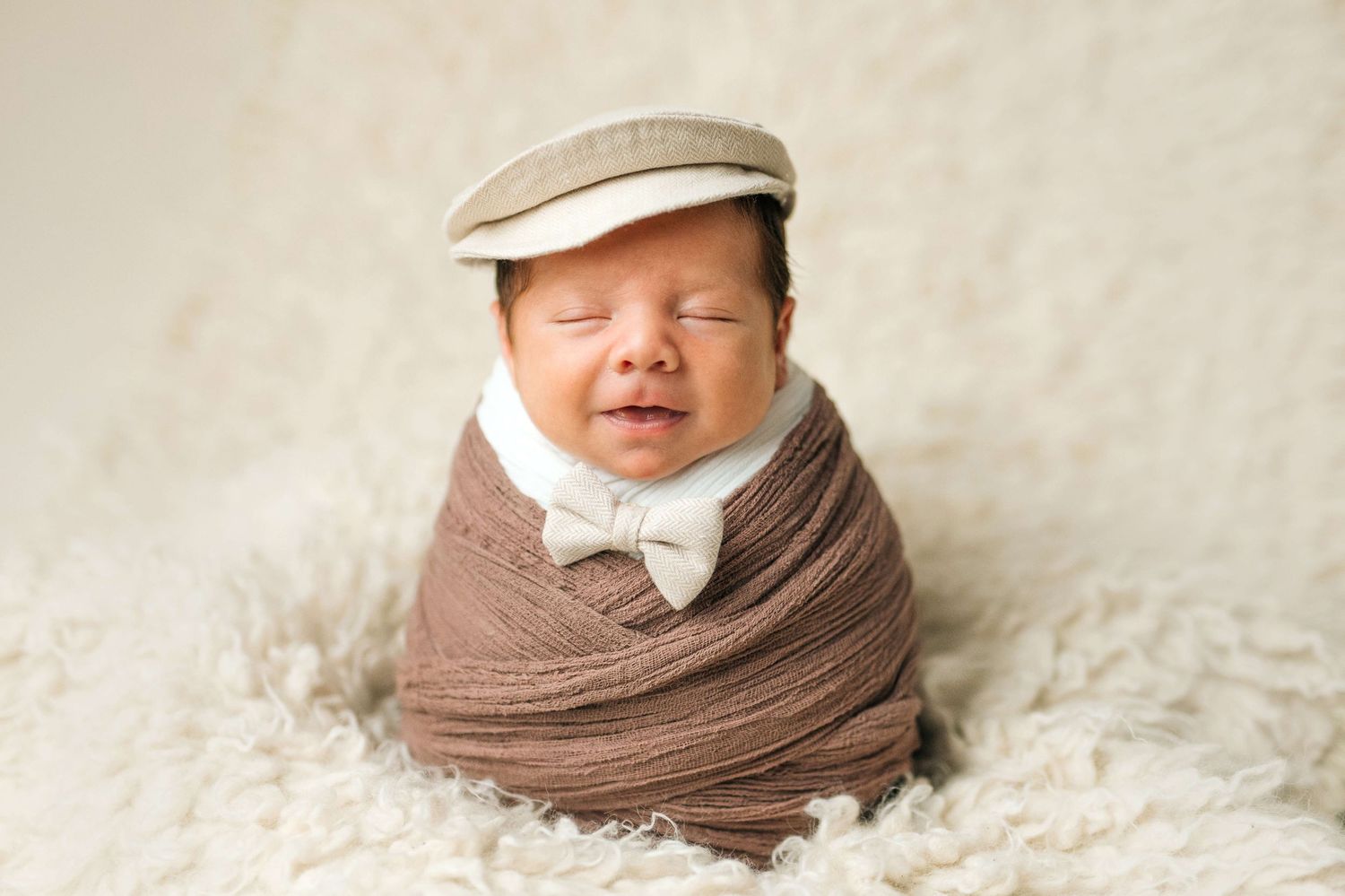 Adorable newborn baby wrapped in brown swaddle wearing a bow tie and flat cap, sleeping peacefully on a fluffy white rug.