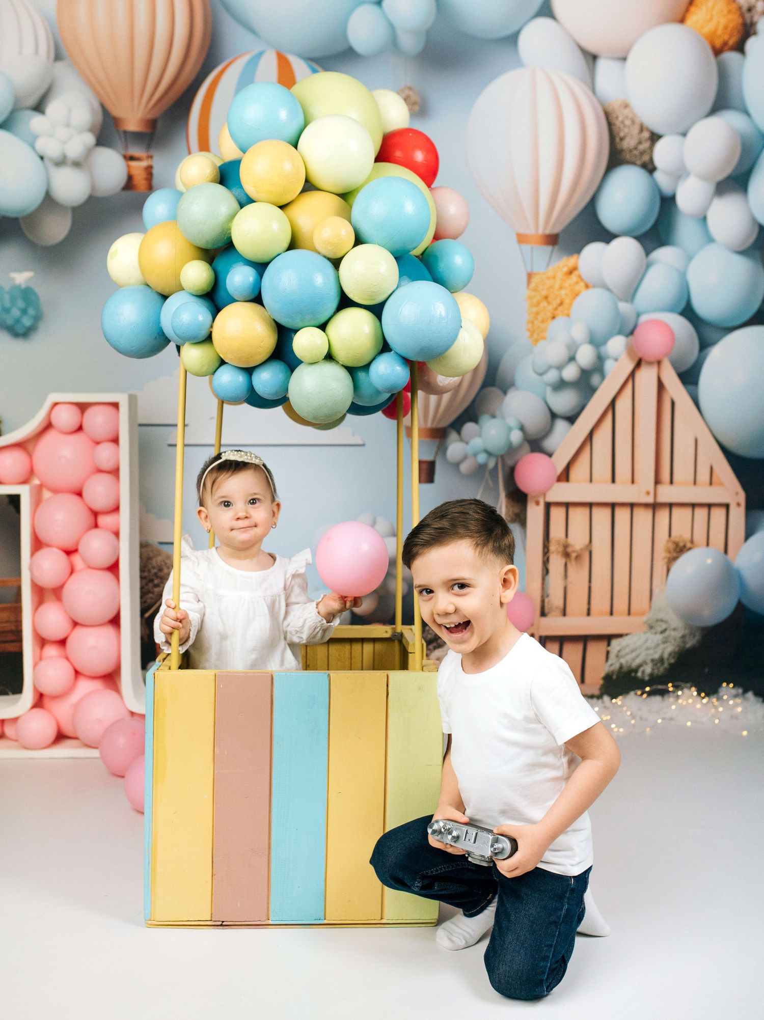 Two children pose in a colorful balloon-themed photo setup with hot air balloon backdrop and festive decorations.