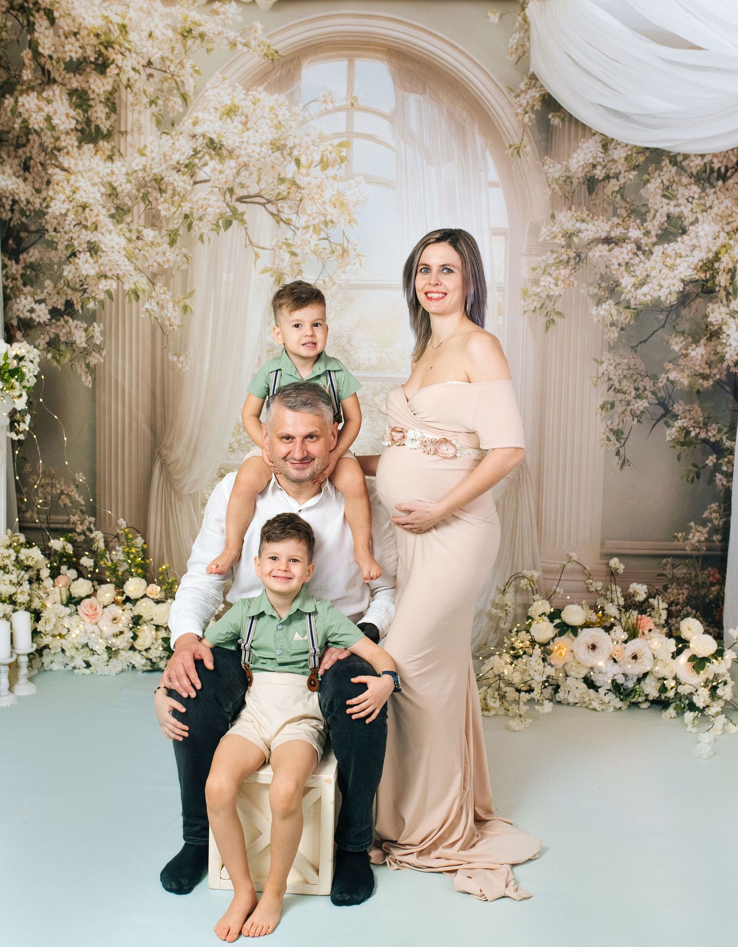 Family maternity photo with pregnant woman in beige gown, man, and two young boys posed among white floral backdrop.