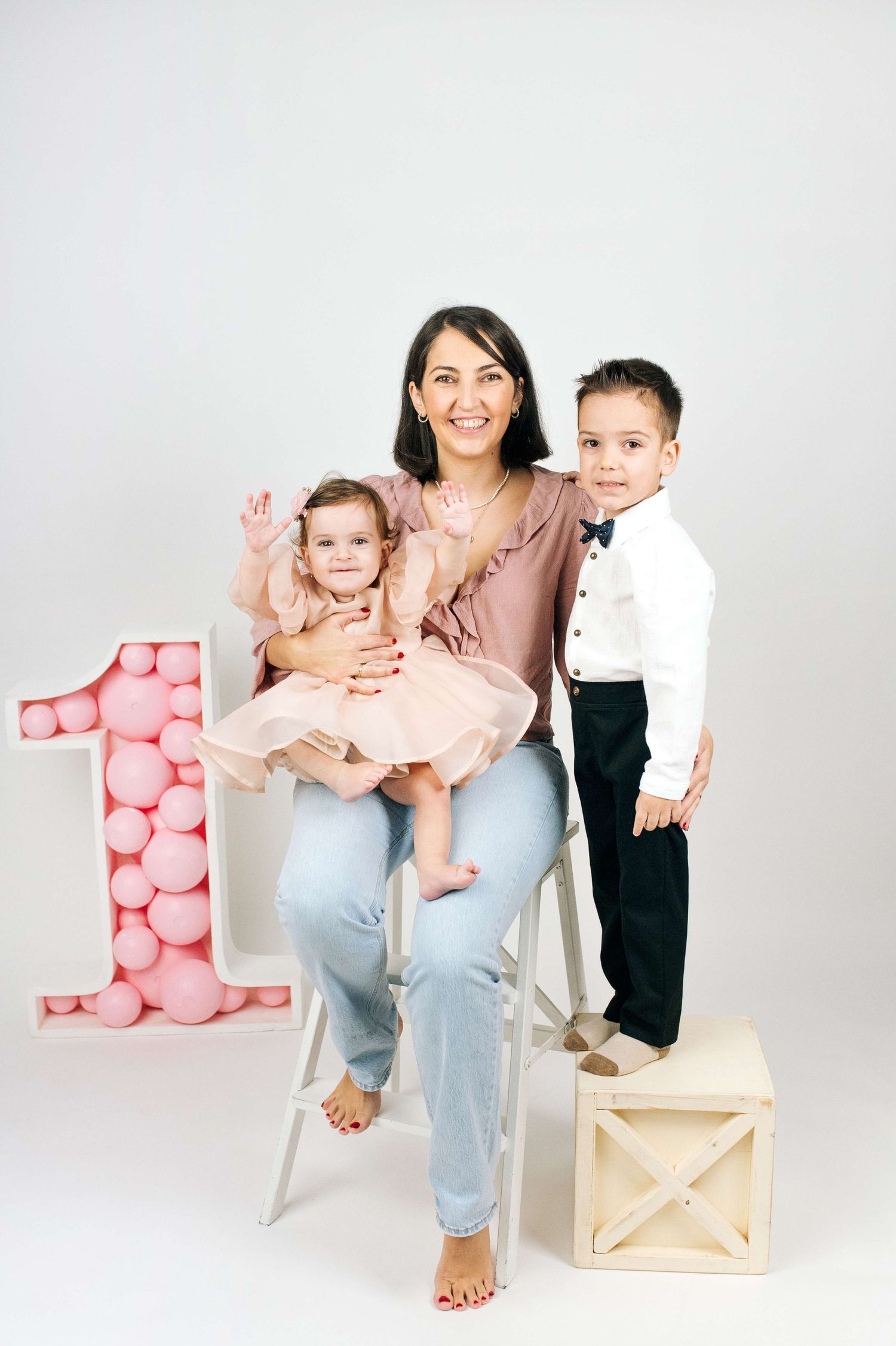 A mother sits on a ladder holding her baby girl in a pink dress, with her young son standing beside her, celebrating a 1st birthday.