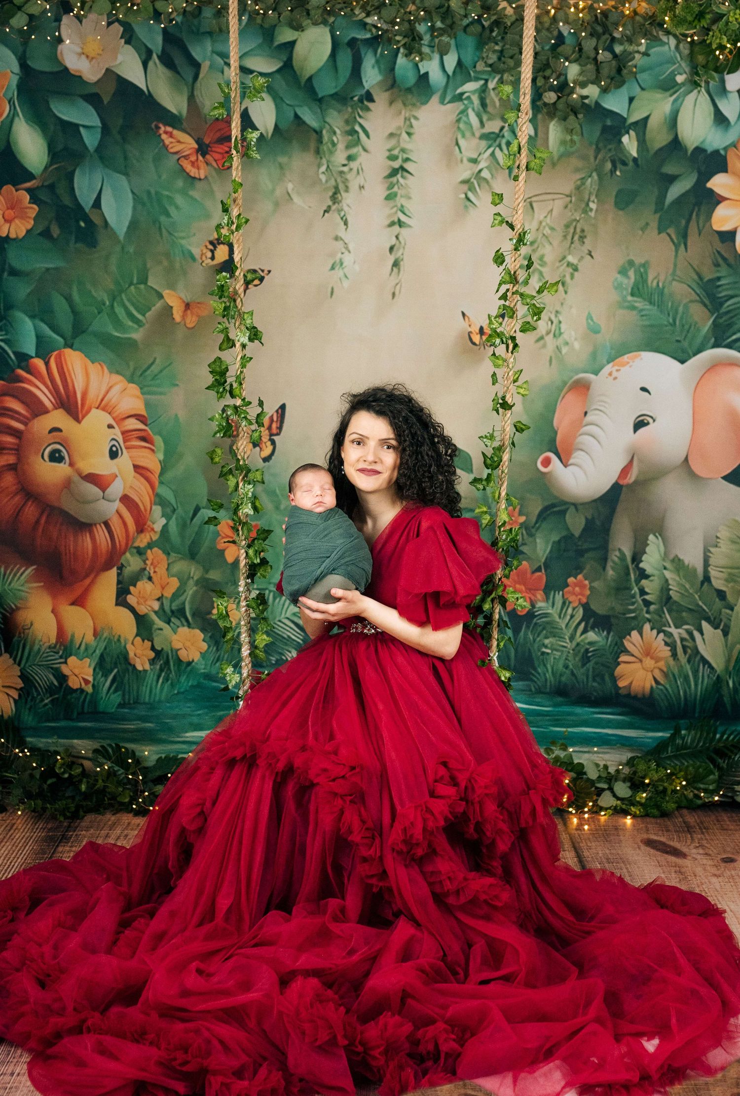 Woman in red gown holds swaddled newborn on jungle-themed swing backdrop with lion and elephant illustrations.