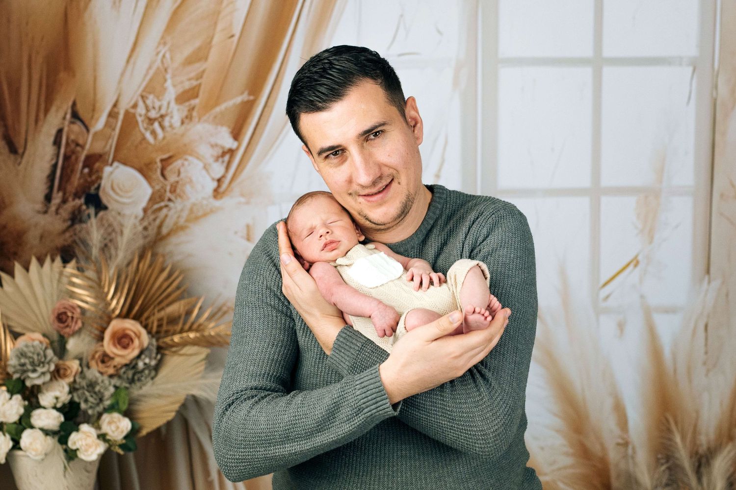 A proud father in a green sweater gently holds his sleeping newborn baby in a warmly decorated studio setting.