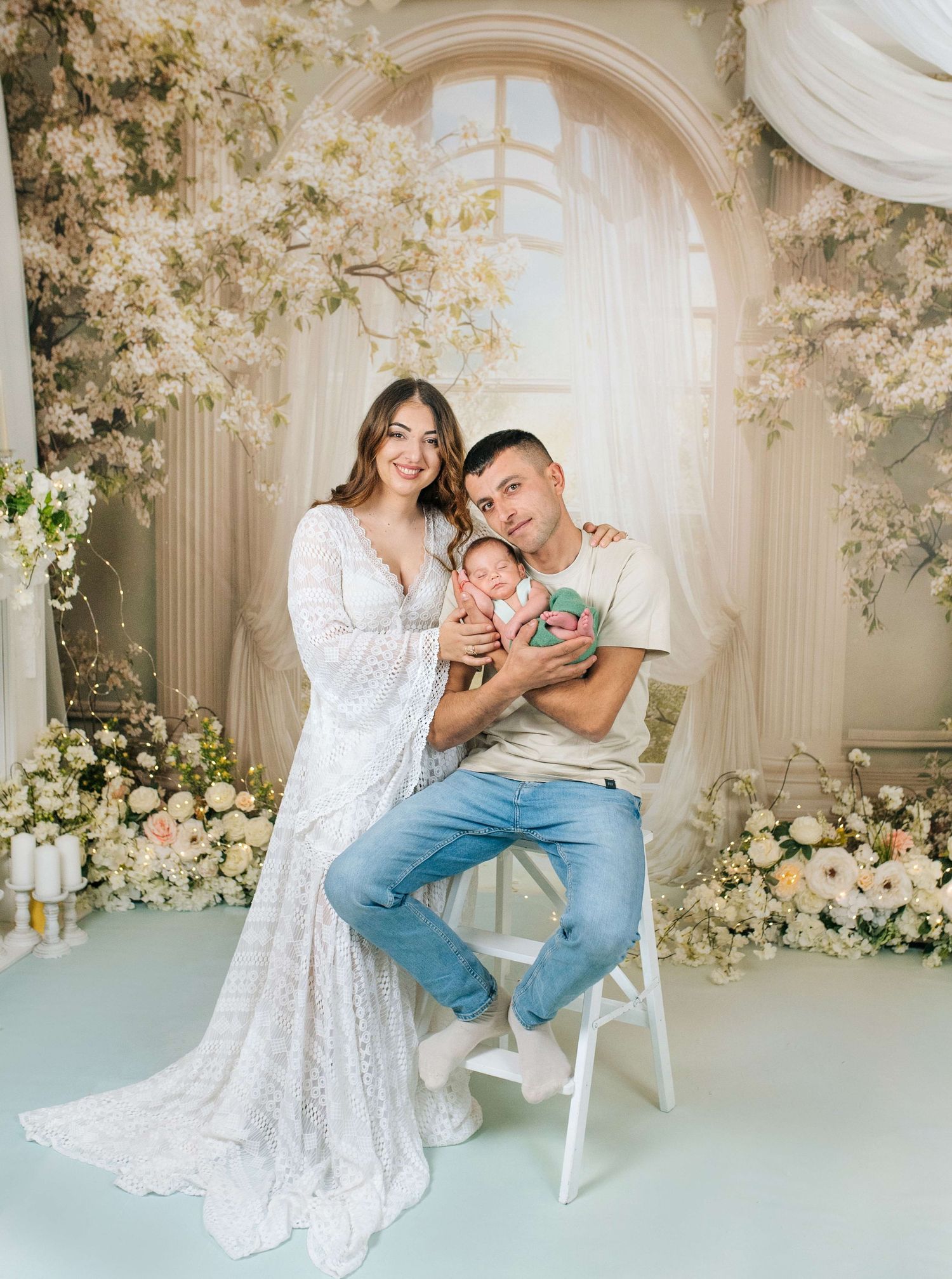A couple in elegant attire poses with their newborn baby amid lush floral decorations in a dreamy studio setting.