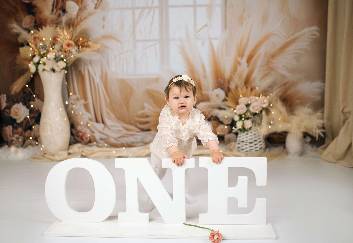 Baby girl in white dress sits behind ONE letters at boho-themed first birthday photo shoot.