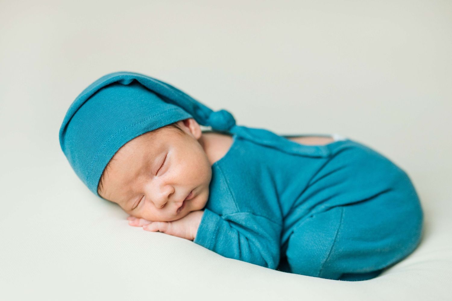 Sleeping newborn baby wrapped in teal swaddle with matching knotted hat on white background.