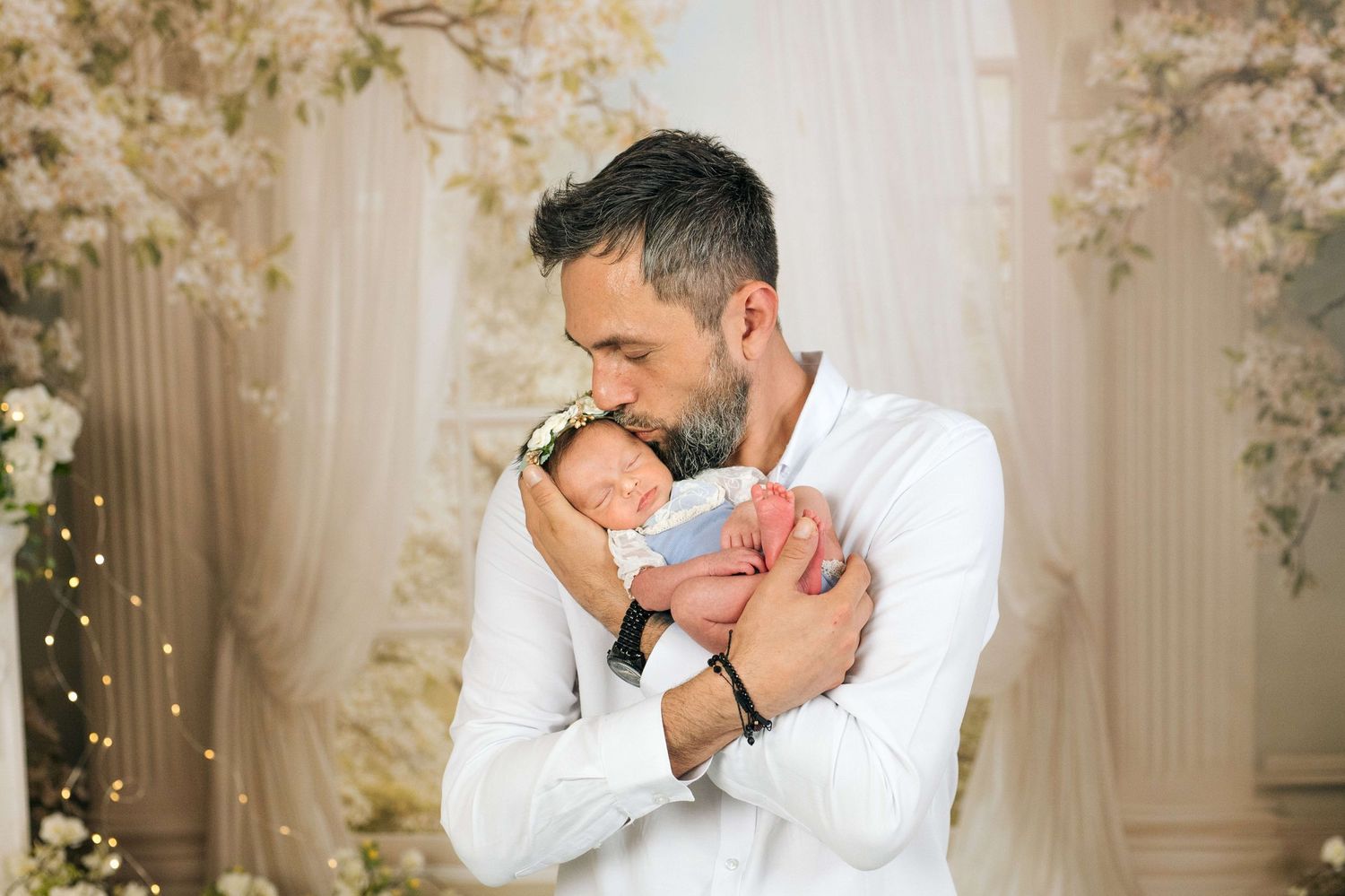 A father in a white shirt tenderly kisses his sleeping newborn baby amid a floral backdrop.