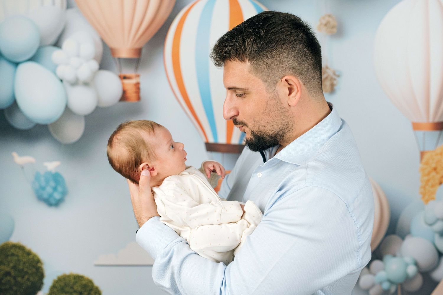 A loving father gently holds his newborn baby in front of a colorful hot air balloon nursery backdrop.