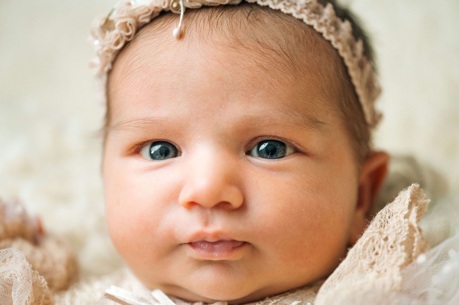 Close-up portrait of a newborn baby with blue eyes wearing a delicate lace headband.