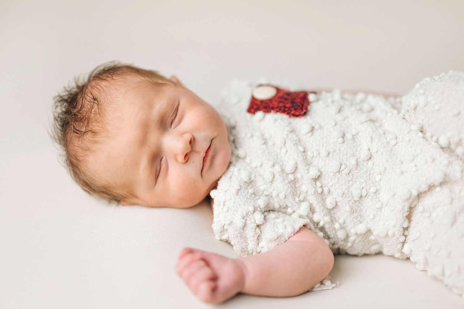 Sleeping newborn baby wrapped in a white textured blanket with a small red accent, lying on a soft white background.