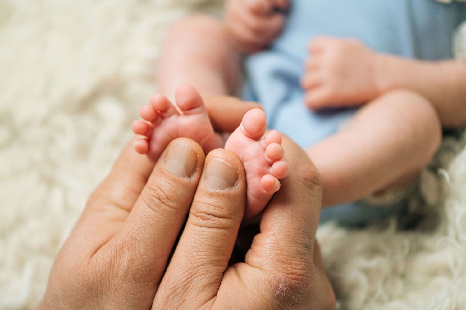 Close-up of adult hands gently holding tiny newborn baby feet on a soft white fluffy blanket.