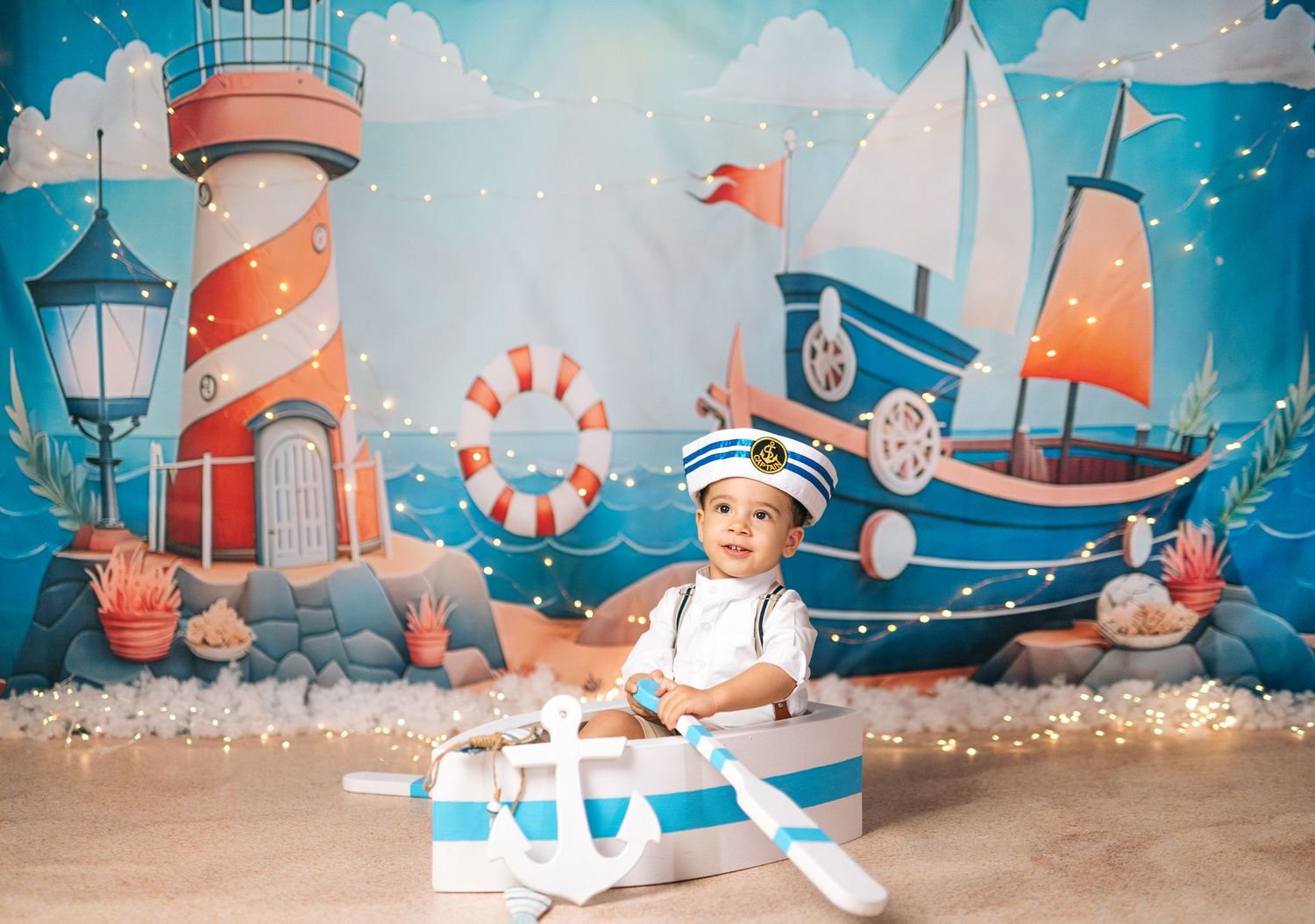 Baby boy in sailor hat sits in decorative boat with nautical backdrop featuring lighthouse and sailboat.