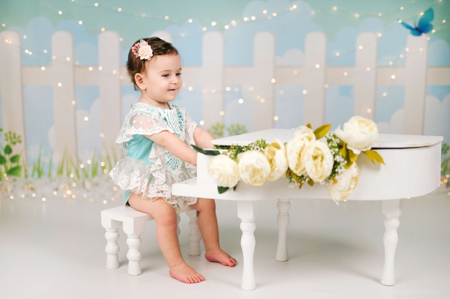 Toddler girl in floral dress sits beside a white mini grand piano with white roses in a whimsical studio setting.