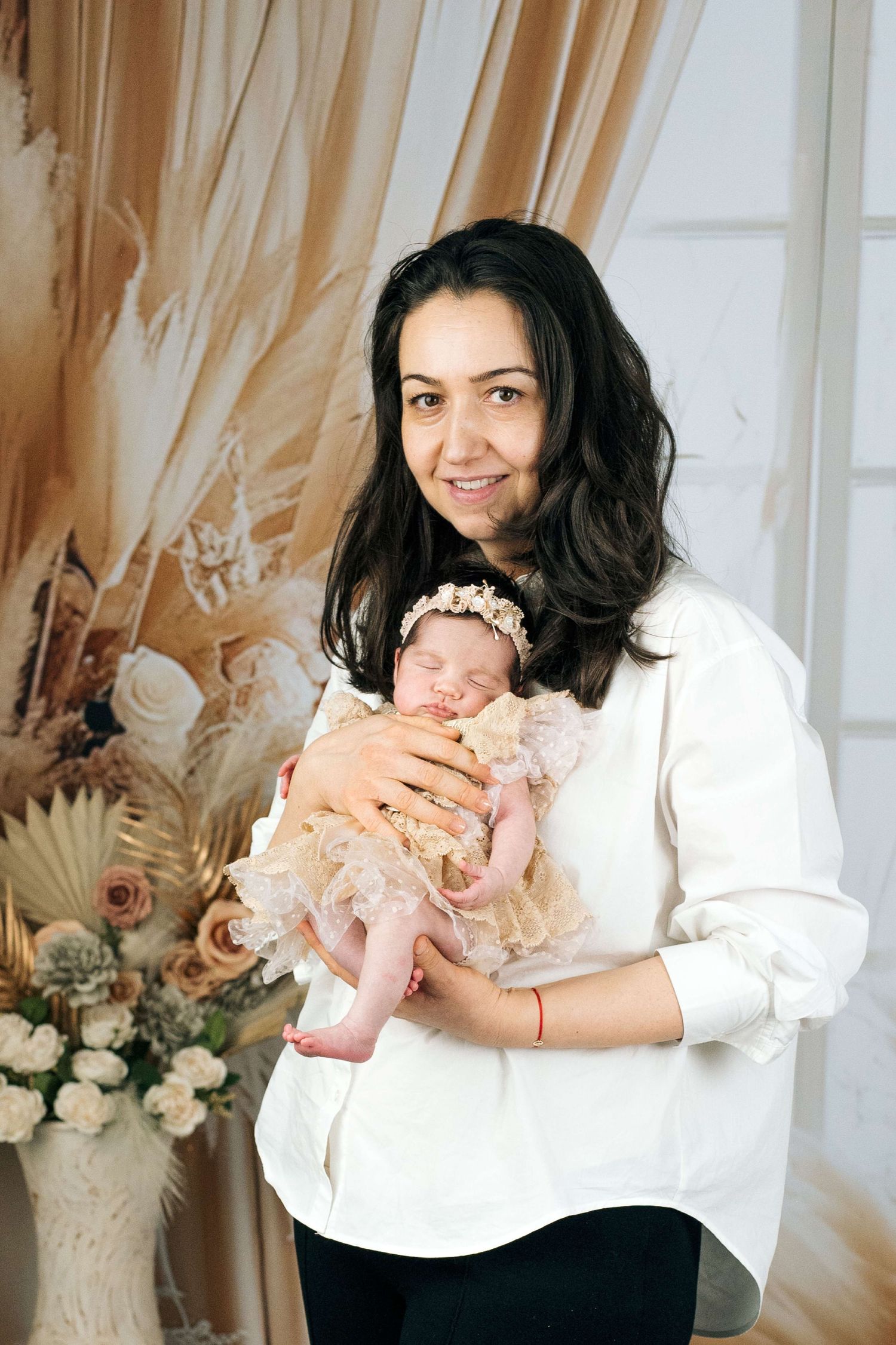 A smiling dark-haired mother in a white blouse holds her sleeping newborn baby girl in a lace dress and floral headband.