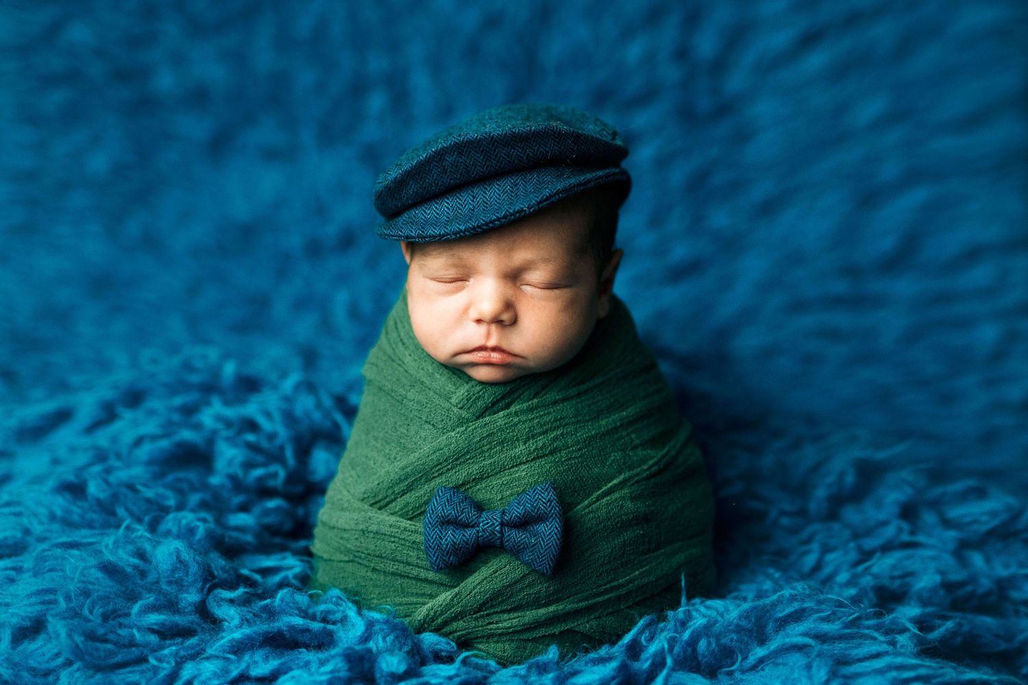 Sleeping newborn baby swaddled in green wrap wearing a navy flat cap and bow tie on blue fluffy backdrop.